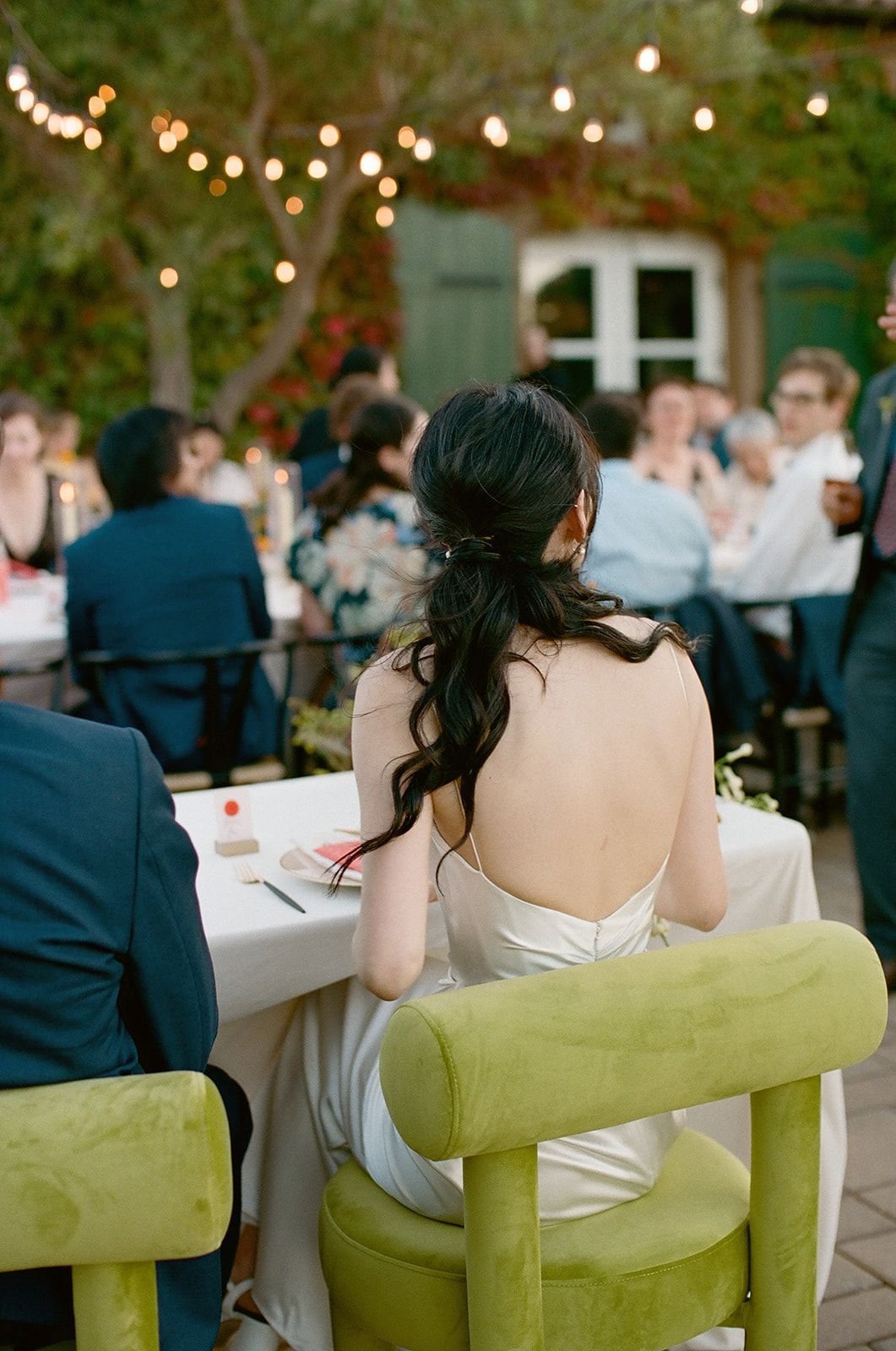 A woman with long dark hair in a white dress sitting at a table during an outdoor event with string lights and guests in the background.