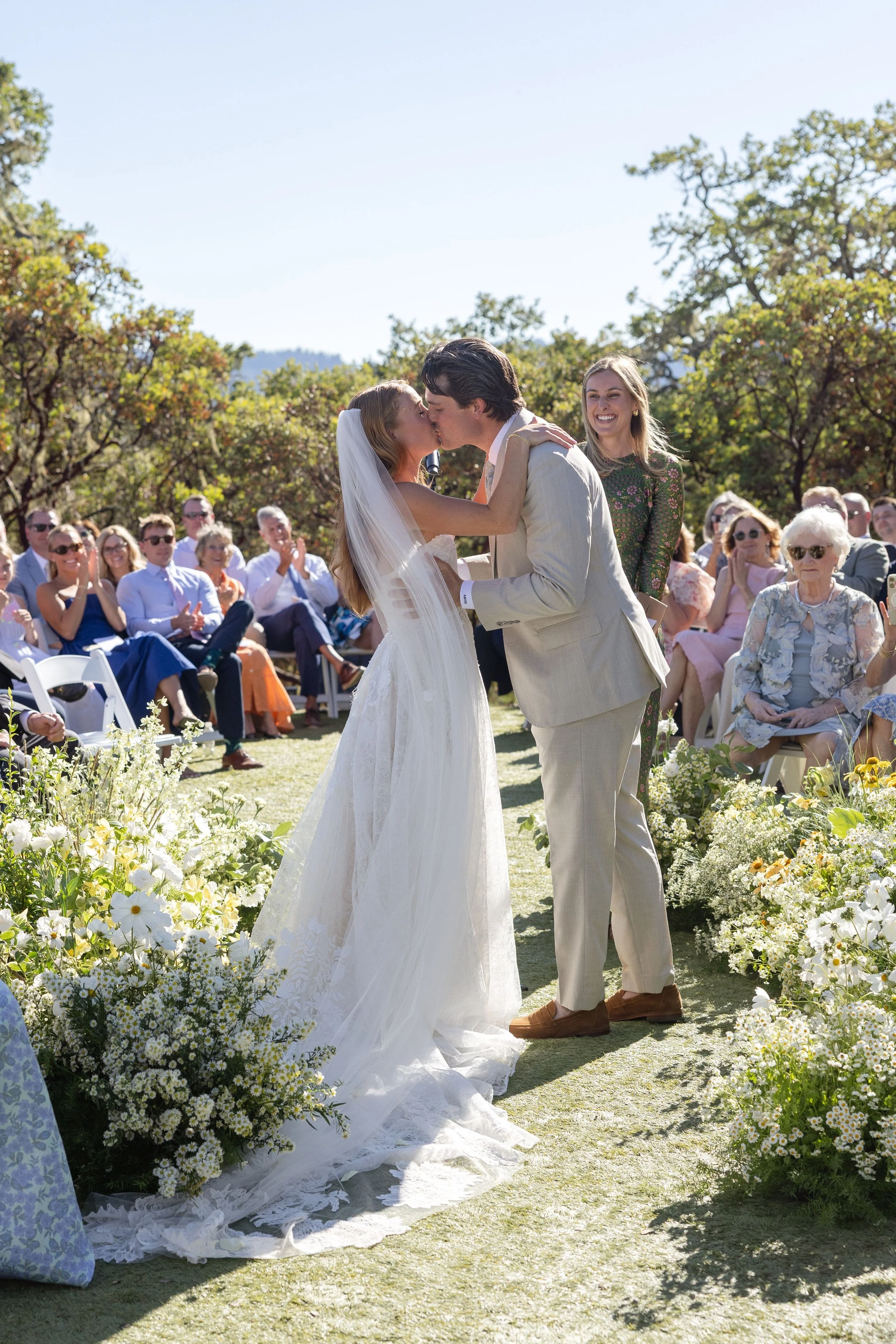 A bride and groom kiss during an outdoor wedding ceremony with guests clapping and smiling in a lush, sunny garden.