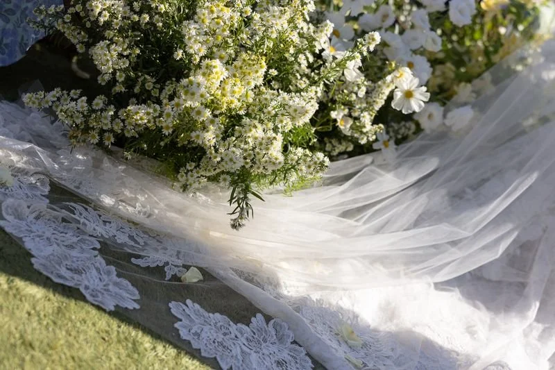Close-up of a bouquet of white flowers, including daisies and small blossoms, on a table decorated with lace and white tulle, outdoors on grass.