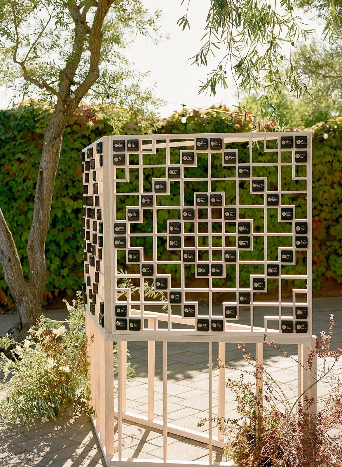 Outdoor installation with a wooden grid featuring numerous small black plaques, set against a background of trees and greenery.