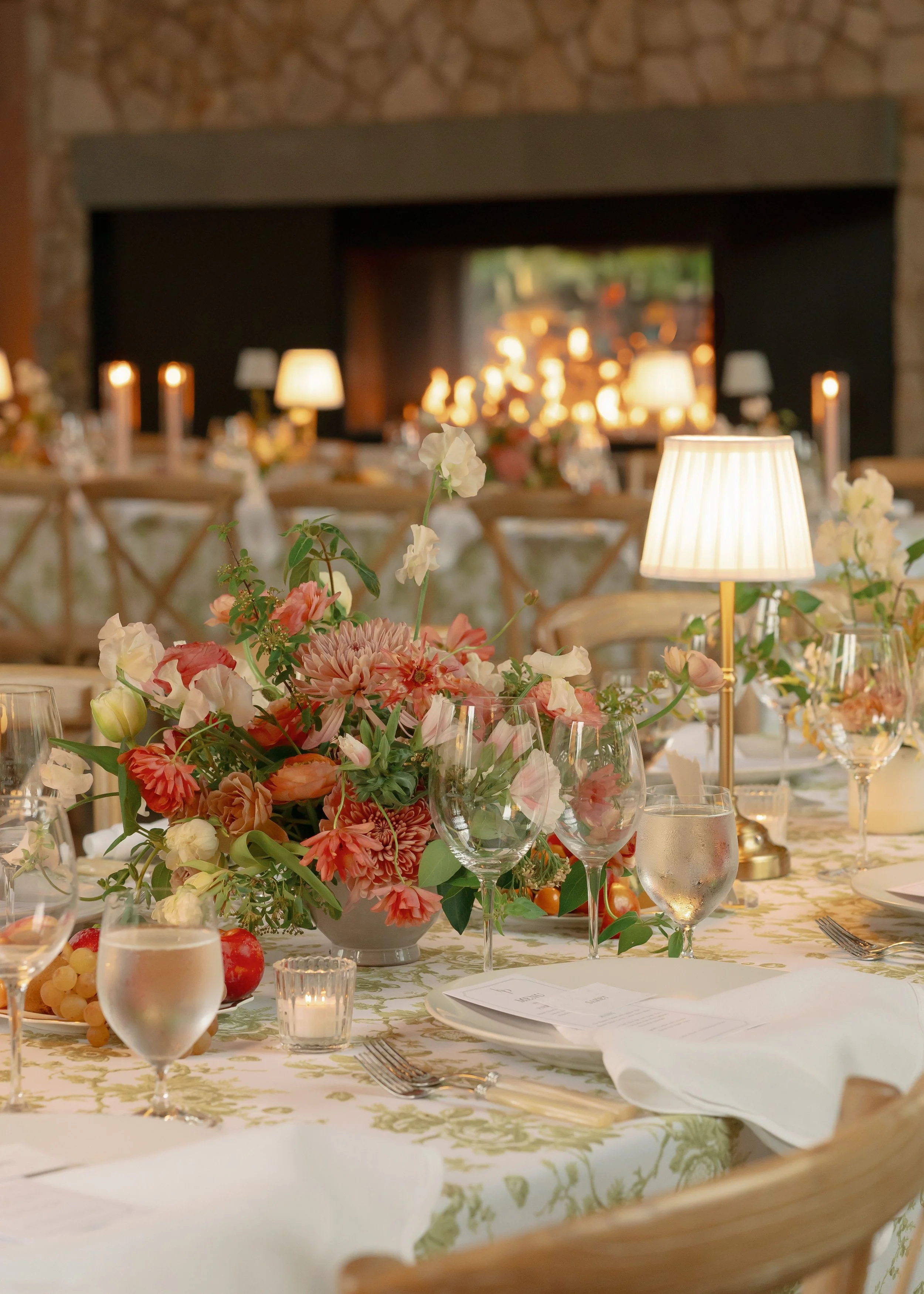 A beautifully decorated dining table with a large floral centerpiece, surrounded by elegant wine glasses, water glasses, candles, and tableware. Soft lighting and a cozy fireplace in the background create a warm atmosphere for a special event or cele
