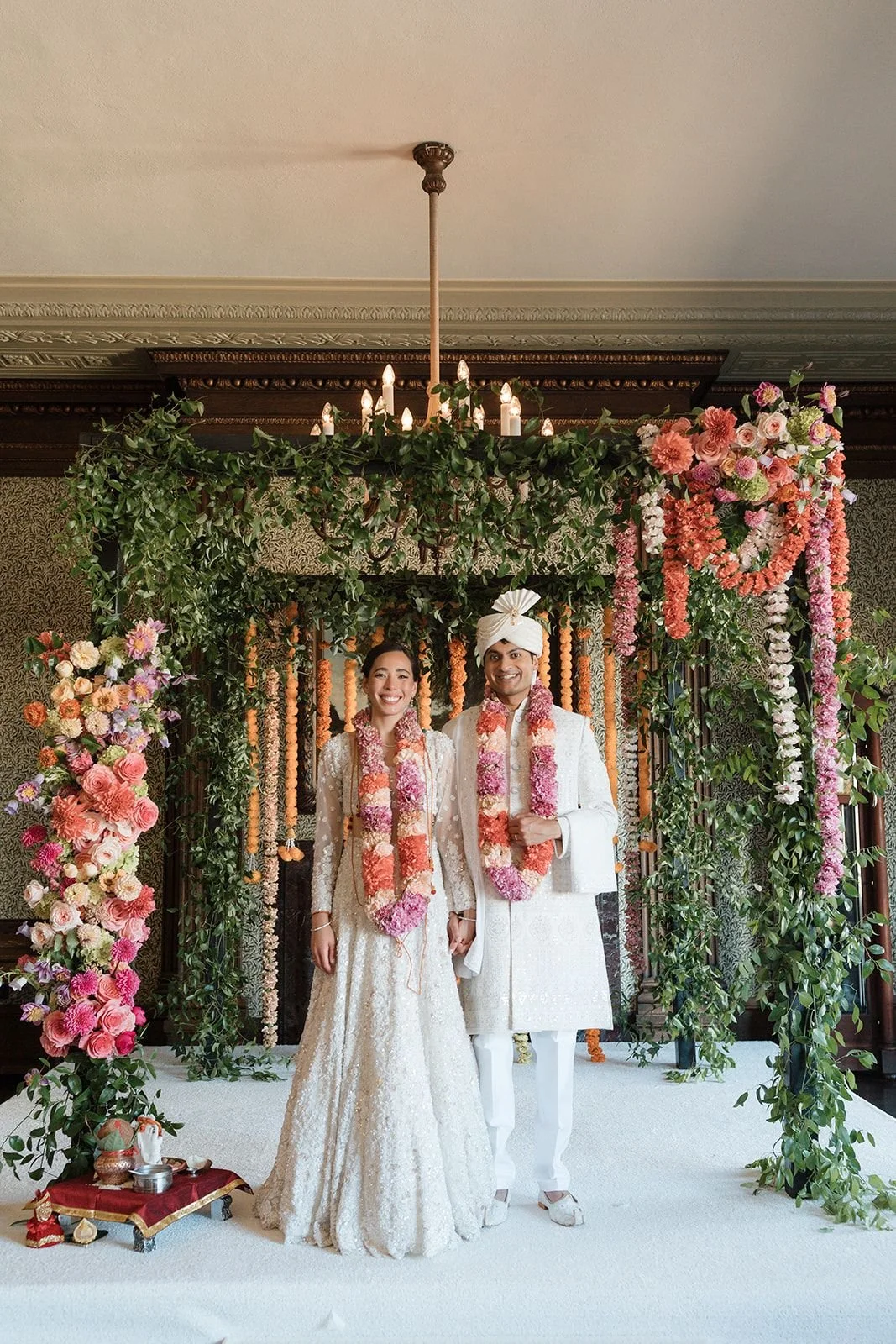 A newlywed Indian couple standing under a floral wedding arch with greenery and pink, peach, and white flowers, in traditional white wedding attire. The bride wears a lace dress with a long train, and the groom wears a sherwani with a turban. Both we