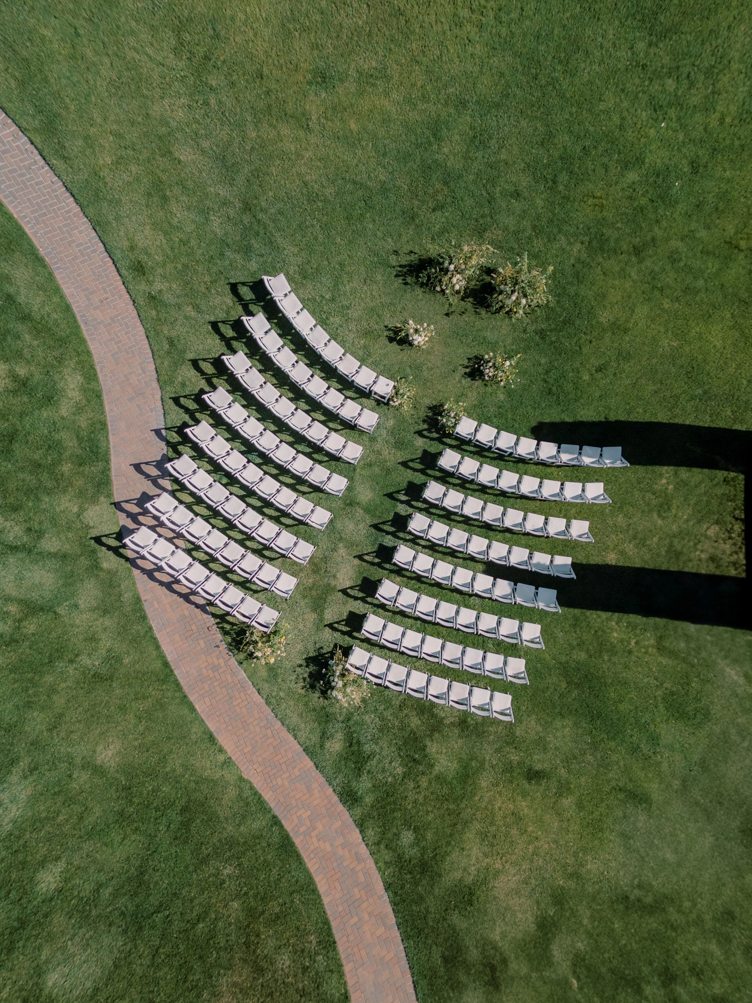 Aerial view of a grassy area with a curved brick pathway on the left and numerous white chairs arranged in curved rows on the right, along with trees and small bushes.