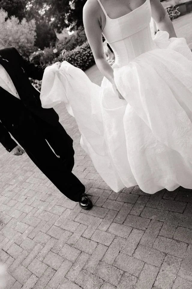 A bride in a white wedding gown walking outdoors on a paved path, holding up her dress slightly, next to a groom in a dark suit.