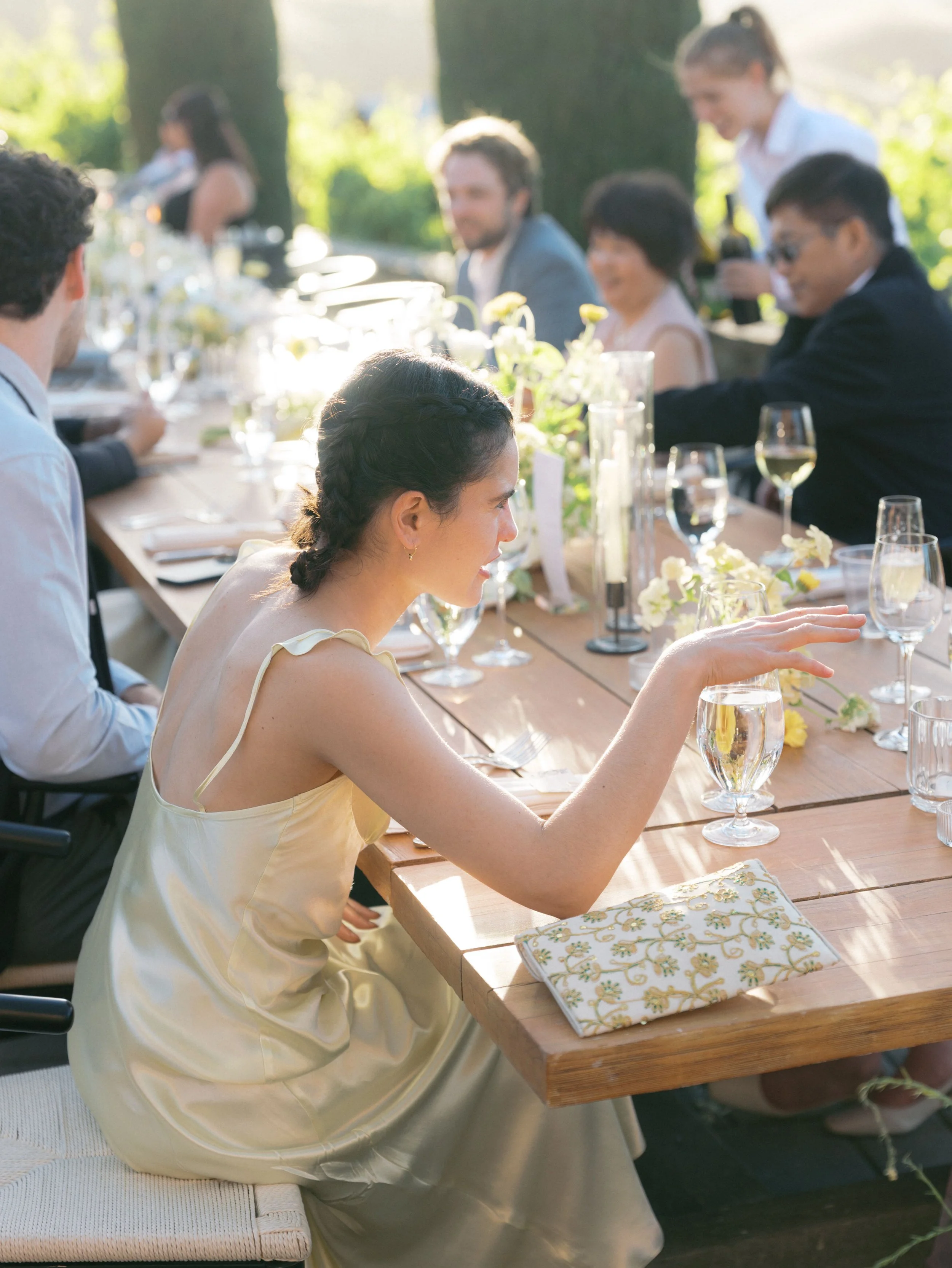 A group of people celebrating at an outdoor wedding or event, sitting at a long table decorated with flowers and drinks, in a sunny garden setting.