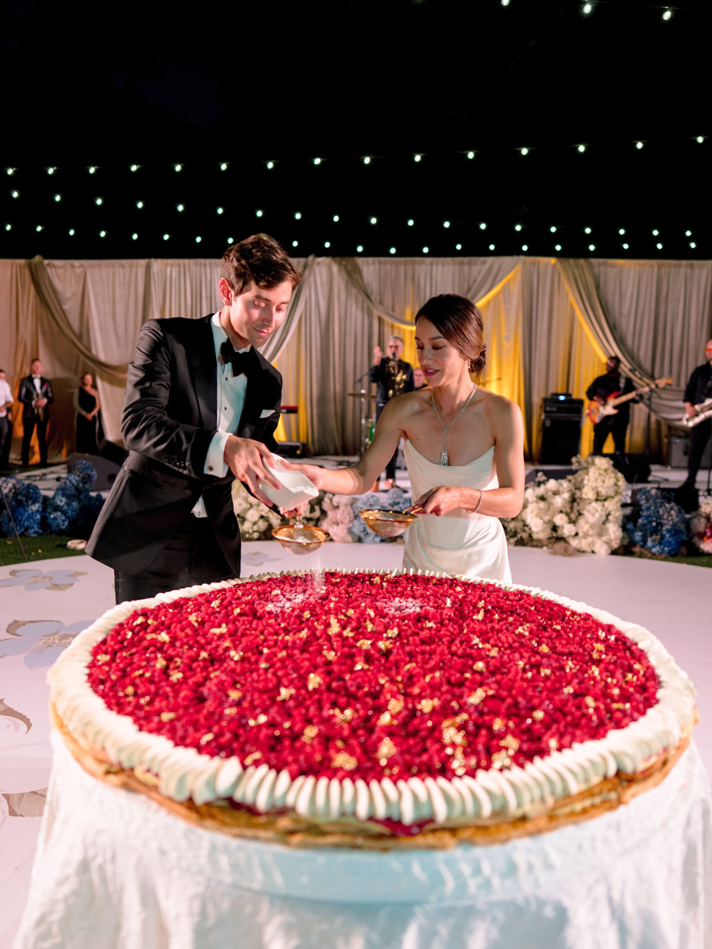 A bride and groom cutting a large berry-topped cake at a wedding reception, with a band playing in the background under string lights.
