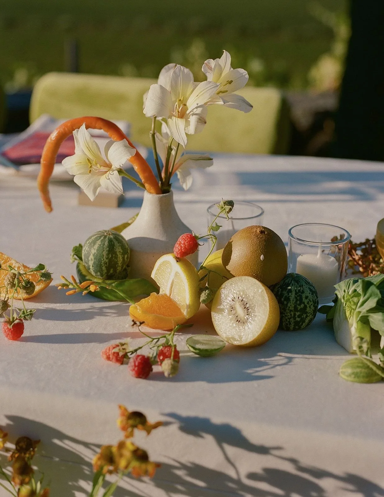 A table set with a white tablecloth, decorated with a white vase holding white lilies, surrounded by sliced citrus fruits, a small watermelon, raspberries, and a candle. The background is out of focus with green trees.