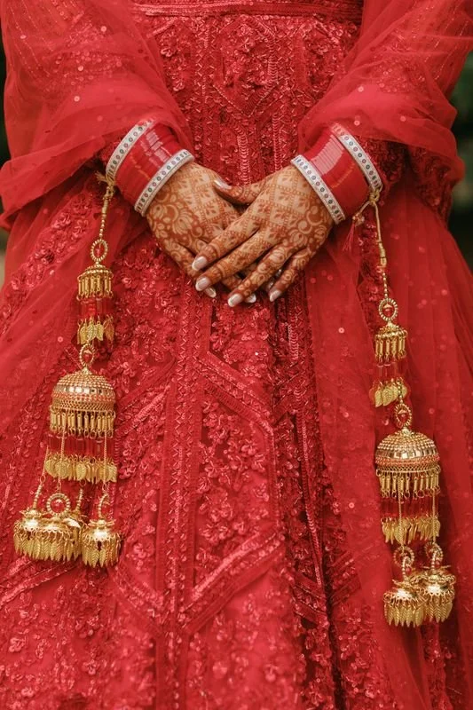 Close-up of hands with henna designs and jewelry, wearing red traditional Indian wedding attire with gold embellishments and tassels.
