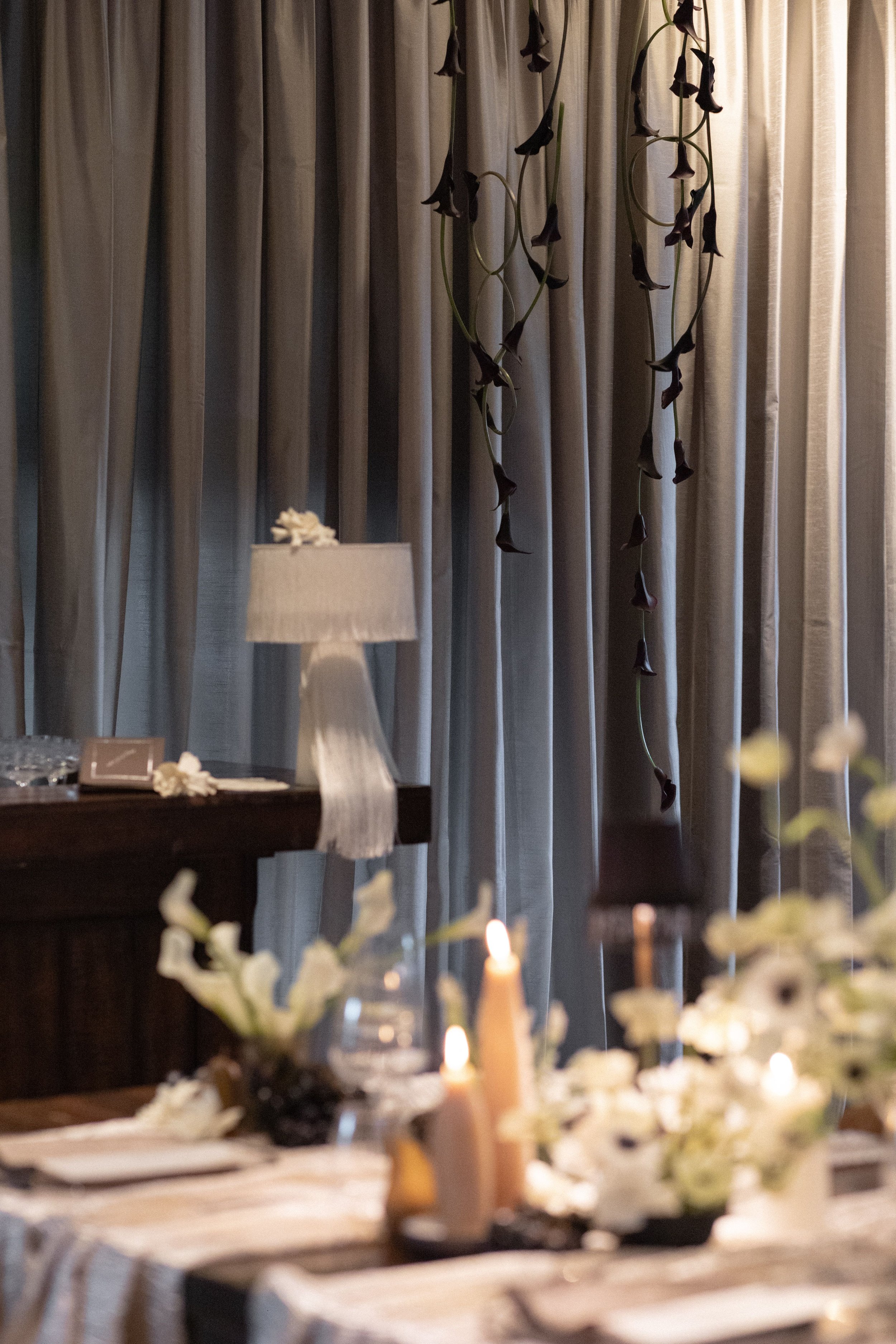Dimly lit interior decor scene with a table set for a celebration, candles and floral arrangements, beige curtains, and purple flowers hanging in the background.