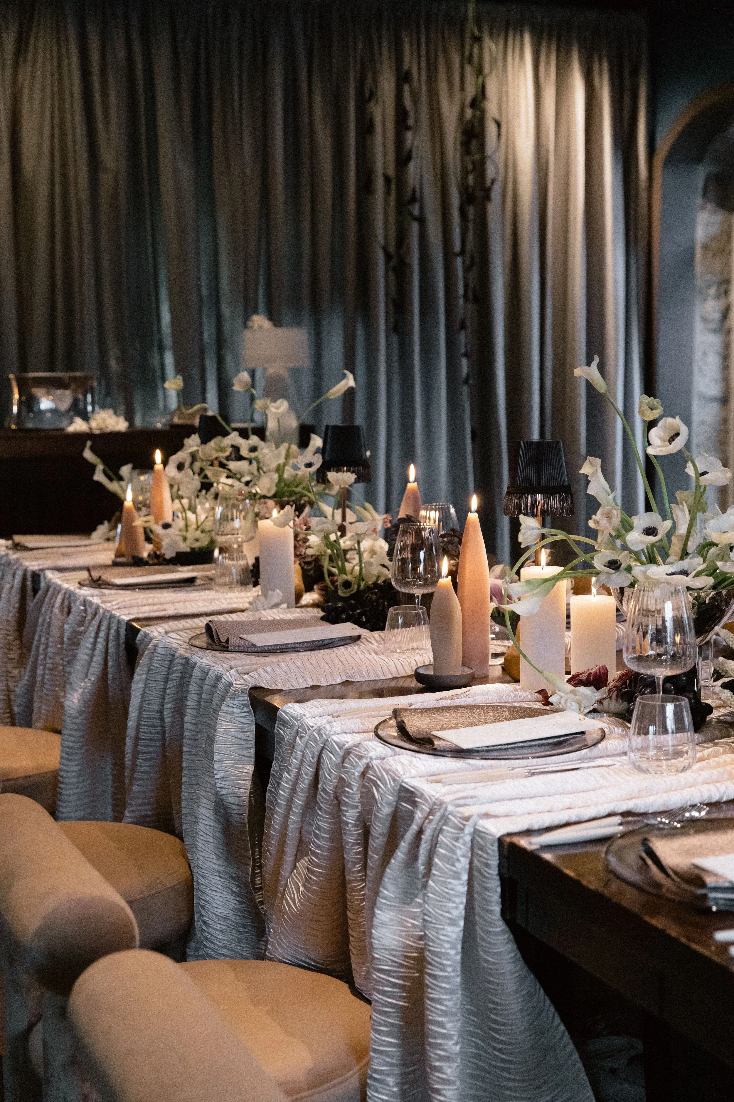 A dining table set for an event with white tablecloths, floral arrangements, candles, and place settings. The background has dark curtains.