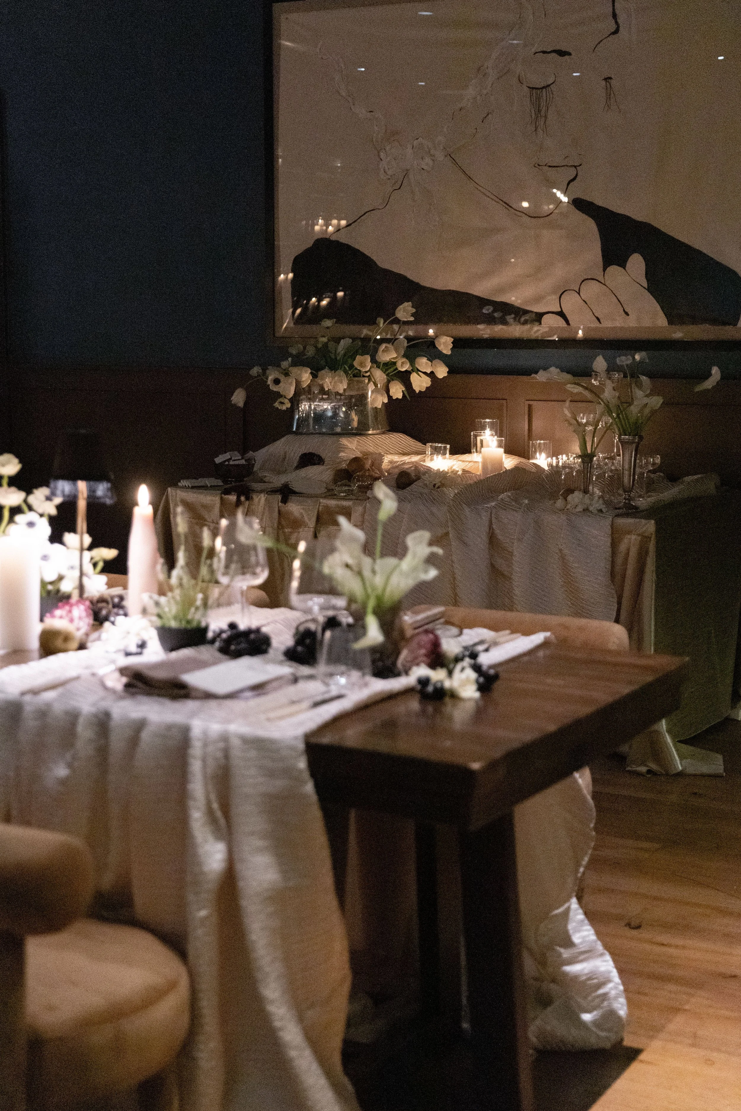 A dimly lit dining room with a table set for a special occasion, decorated with white flowers and candles. There is a large, abstract black and white line art portrait of a woman hanging on the wall. The table has white linen, plates, napkins, and gl