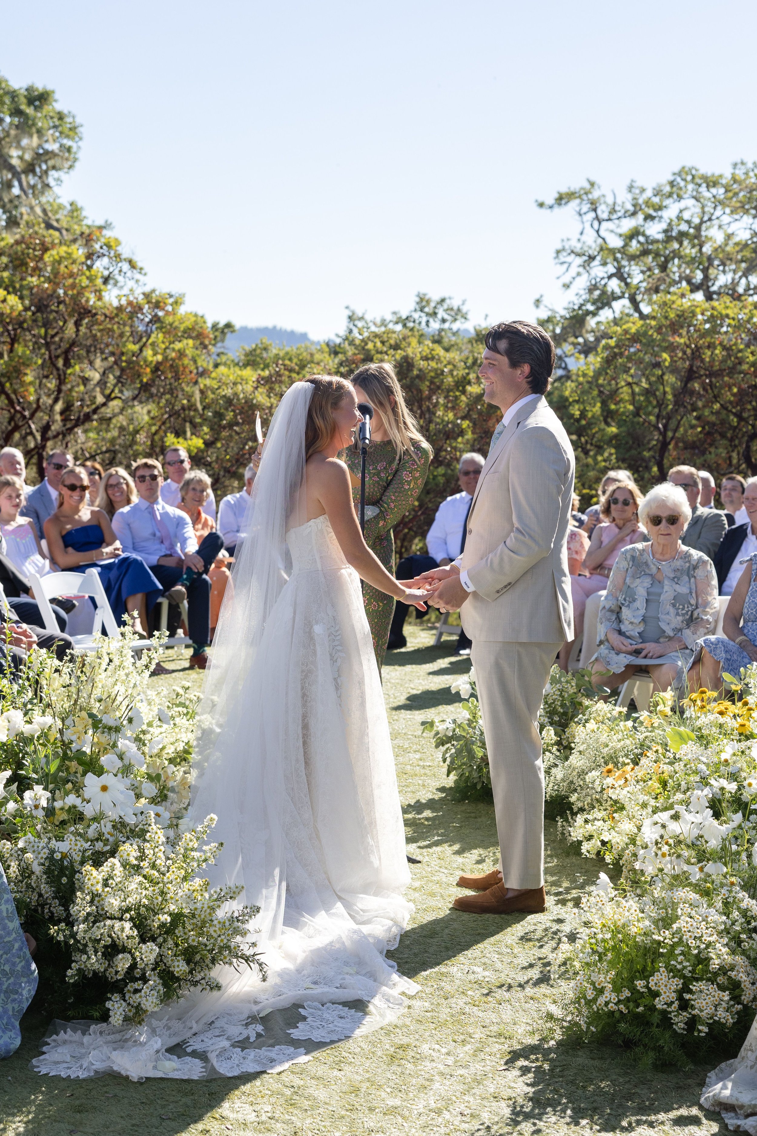 A wedding ceremony outdoors with a bride and groom holding hands and smiling at each other, surrounded by guests and white flowers.