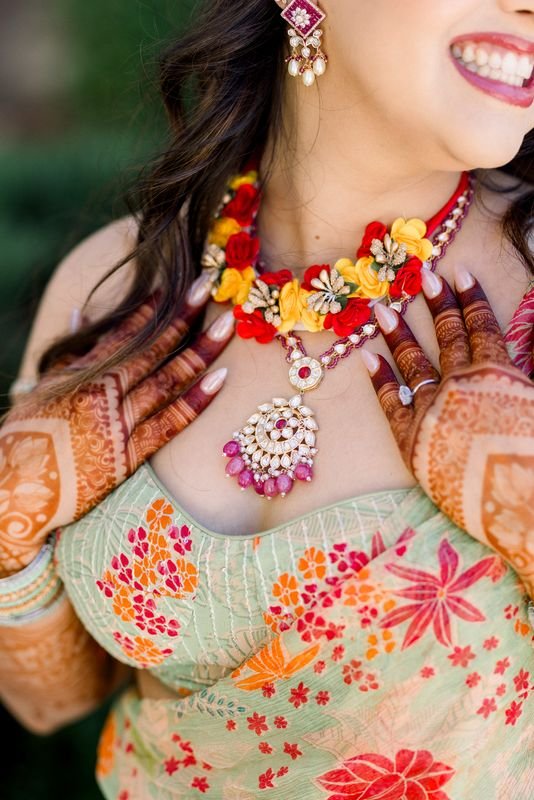 Close-up of a woman wearing colorful floral jewelry, pastel green dress with red and orange floral patterns, henna on hands, smiling, with part of her face visible.