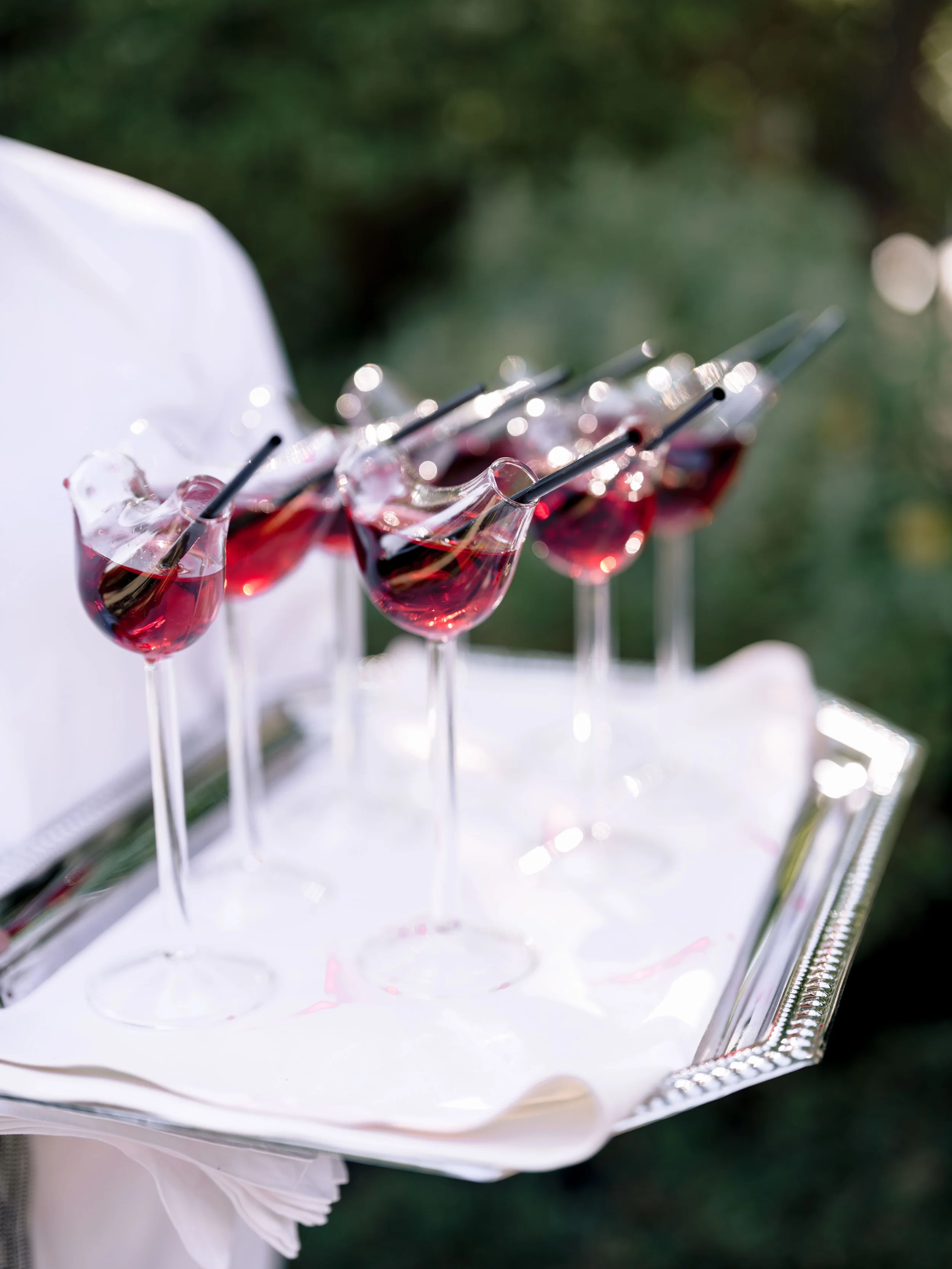 A silver tray holding several glasses of red wine with black straws, with a person in white attire holding the tray outdoors.
