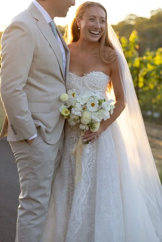 A bride in a white lace wedding dress holding a bouquet of white and yellow flowers, smiling at a groom in a beige suit outdoors during sunset.