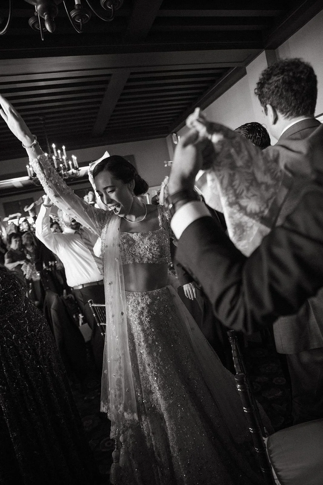 A bride dancing and smiling at her wedding reception, surrounded by guests in formal attire, in a black and white photo.