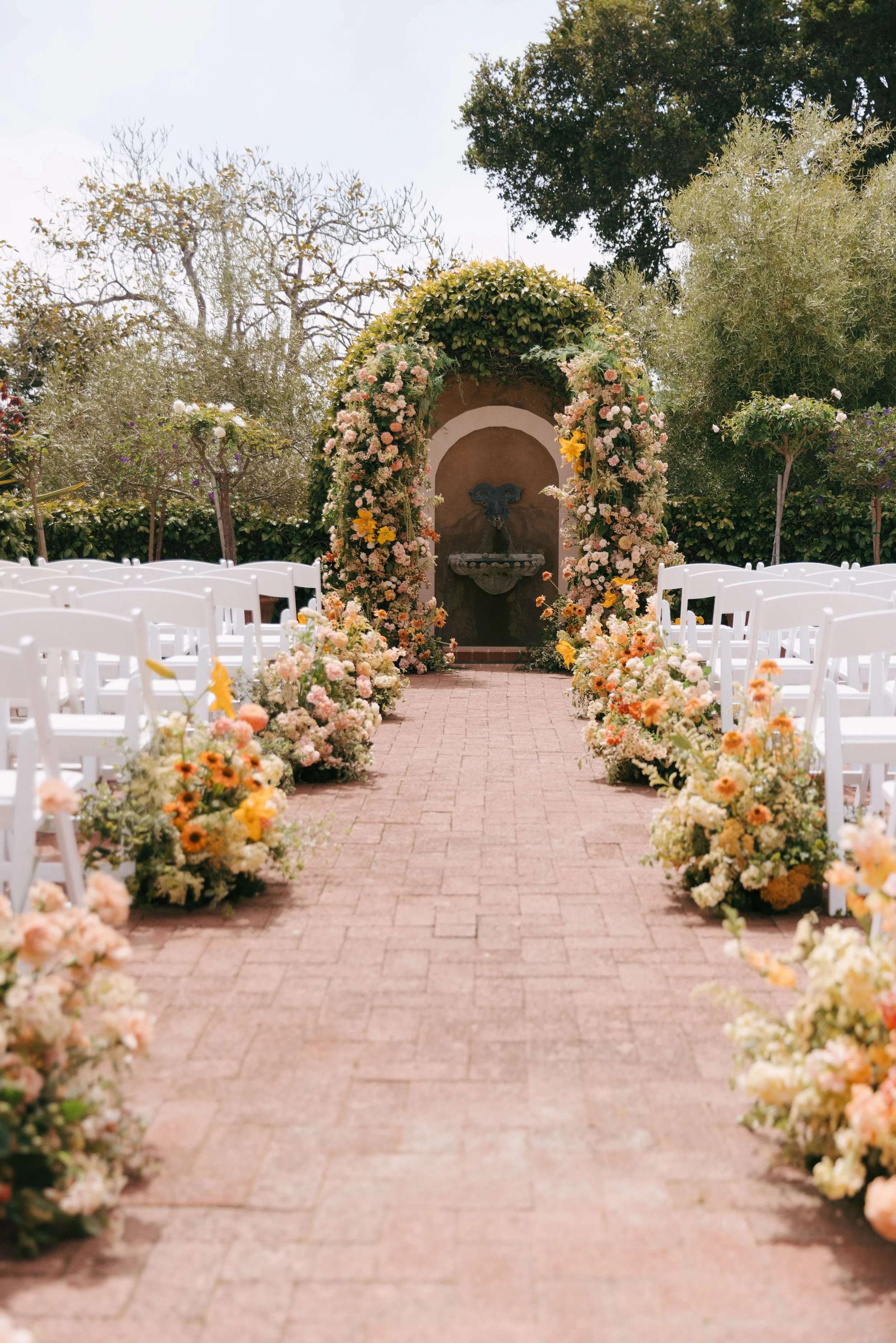 Outdoor wedding ceremony setup with white chairs on either side of a brick aisle, decorated with colorful flowers, leading to a stone fountain adorned with flowers and greenery under an arch, surrounded by trees.