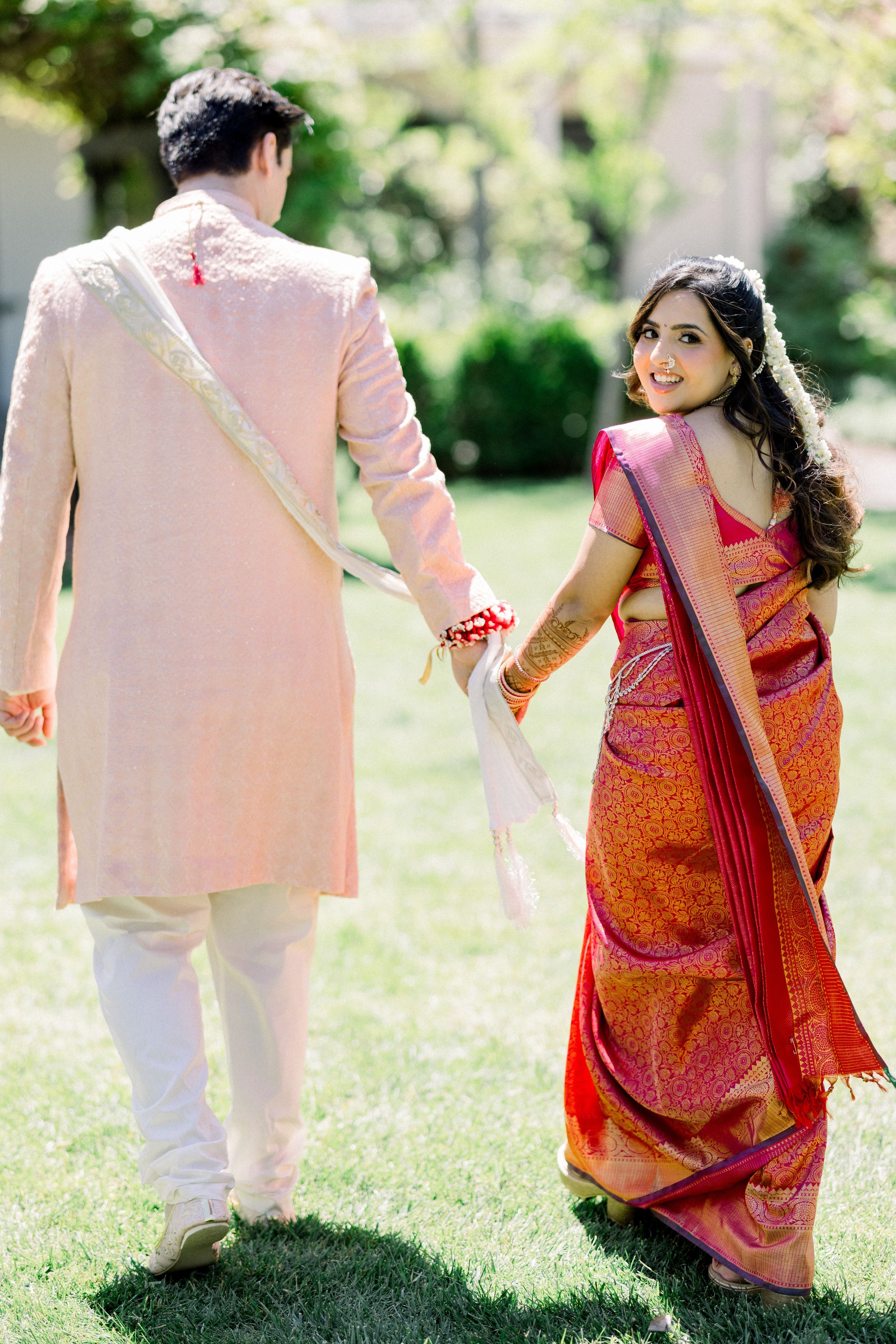 A couple dressed in traditional Indian attire holding hands outdoors in a garden with green trees and grass, during daytime.