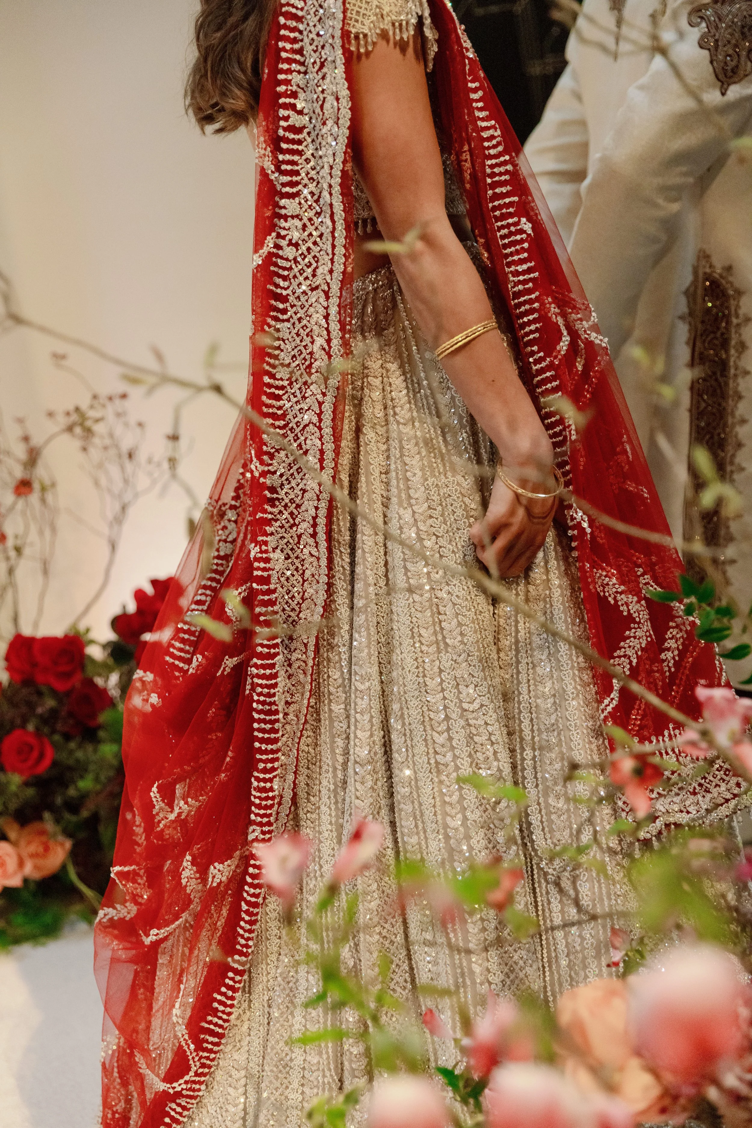 Close-up view of a woman dressed in a traditional Indian bridal outfit with intricate gold and silver embroidery, wearing a red and gold dupatta, gold bangles, and a heavily embellished lehenga, surrounded by floral decorations with roses and blossom