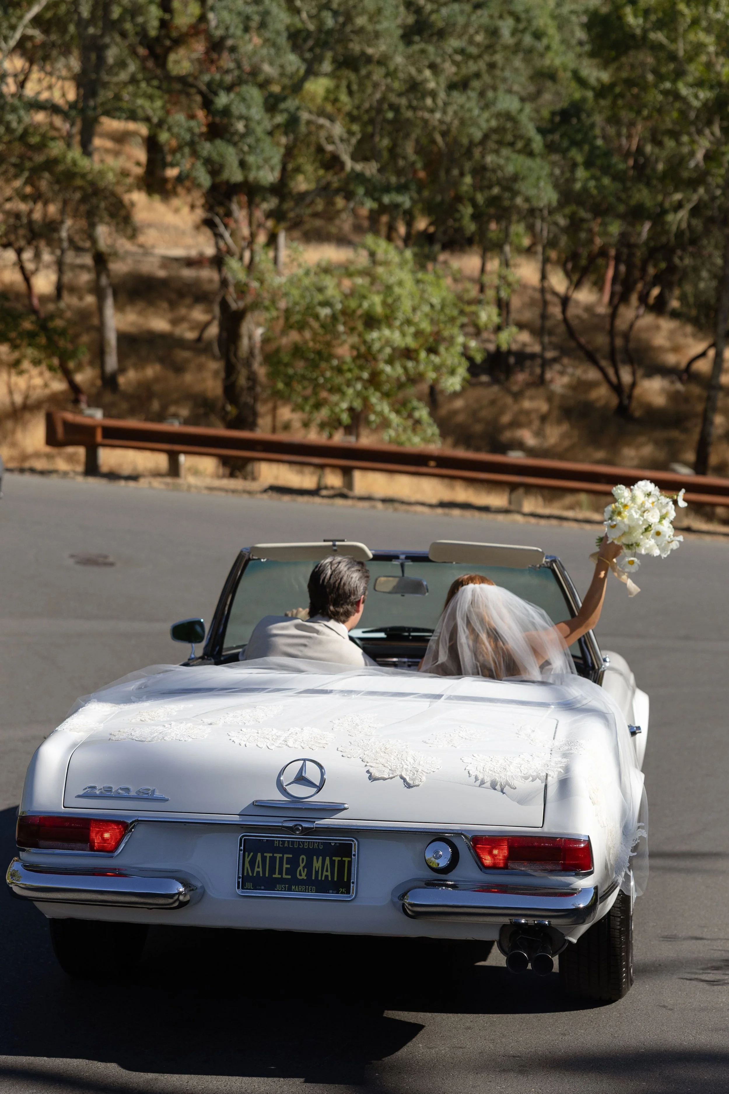 A white convertible Mercedes-Benz with a wedding bride and groom inside, with the bride holding a bouquet of white flowers, on the road with a background of trees and a guardrail.