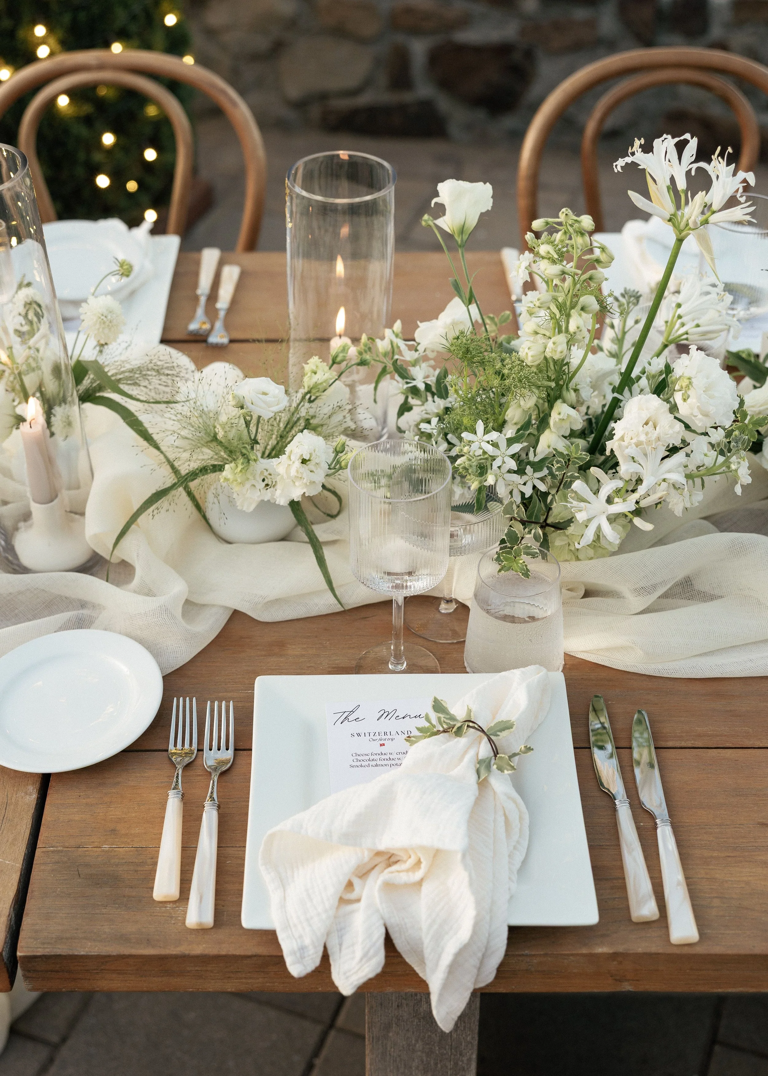 A decorated dining table with white flowers, candles, glasses, and cutlery on a wooden table, set for a meal.