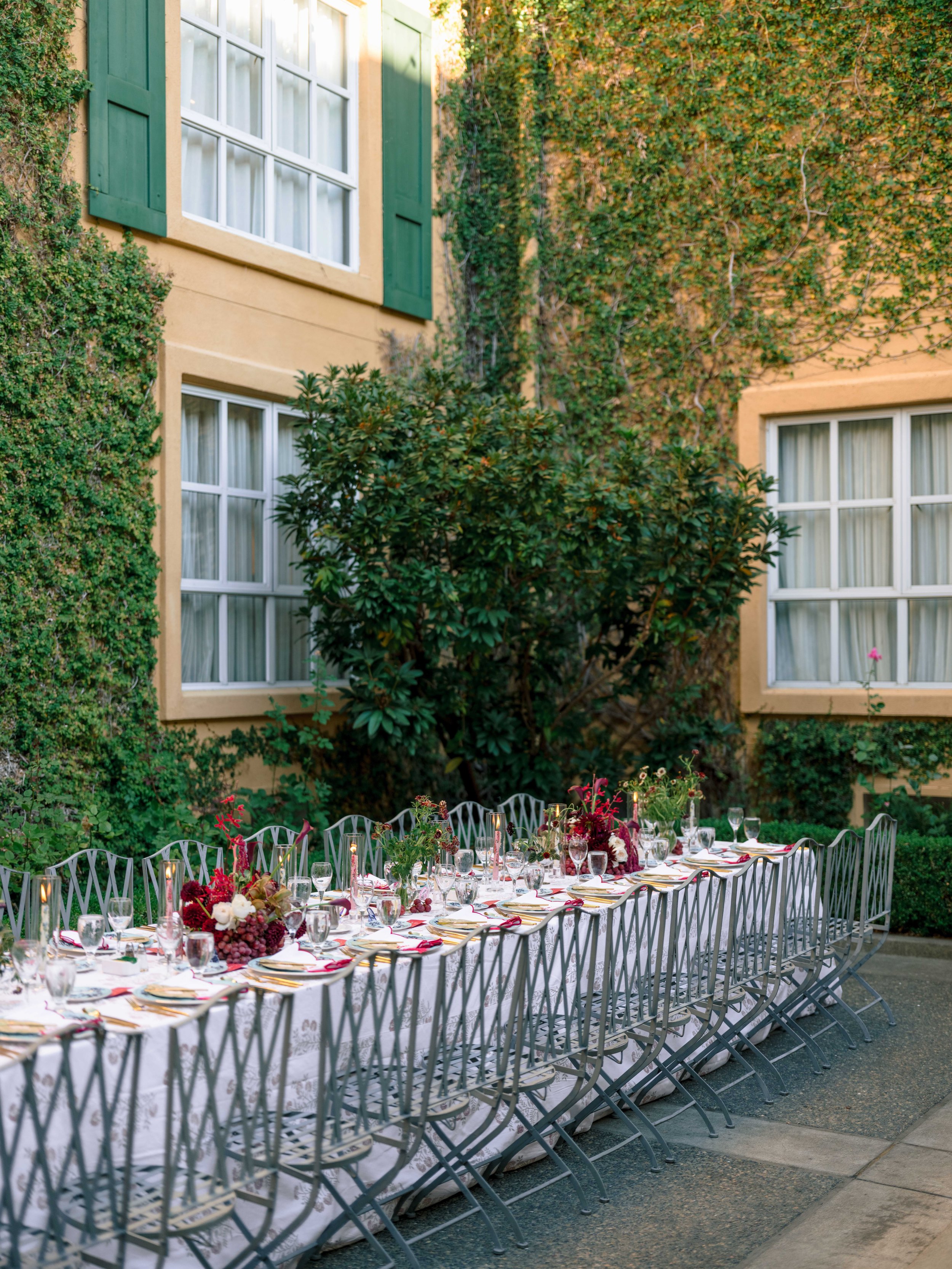 An outdoor banquet table set with floral centerpieces, glassware, and tableware, arranged in front of a building with ivy-covered walls, large windows, and green shutters.