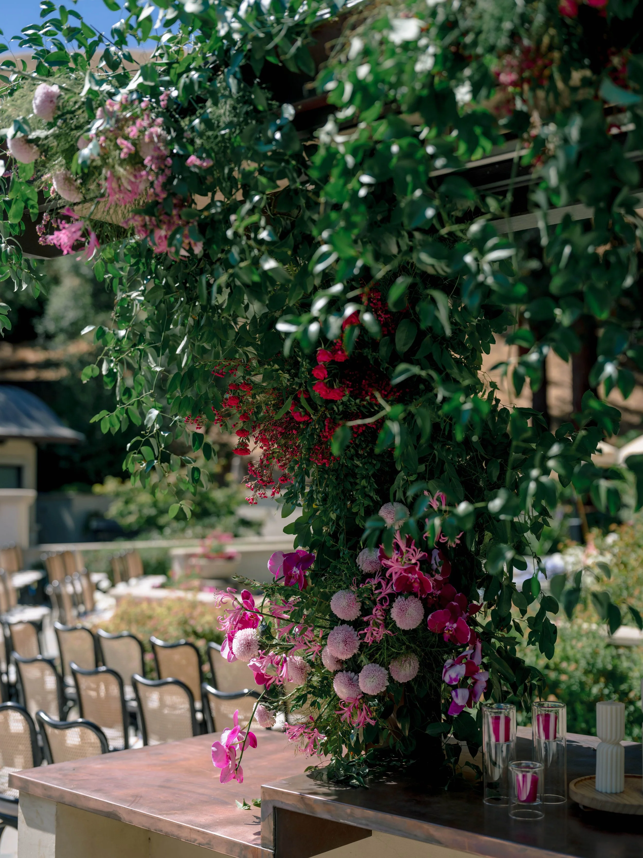 A large floral arrangement on a table with pink, white, and purple flowers, set in an outdoor area with chairs and a building in the background, likely for a special event or celebration.