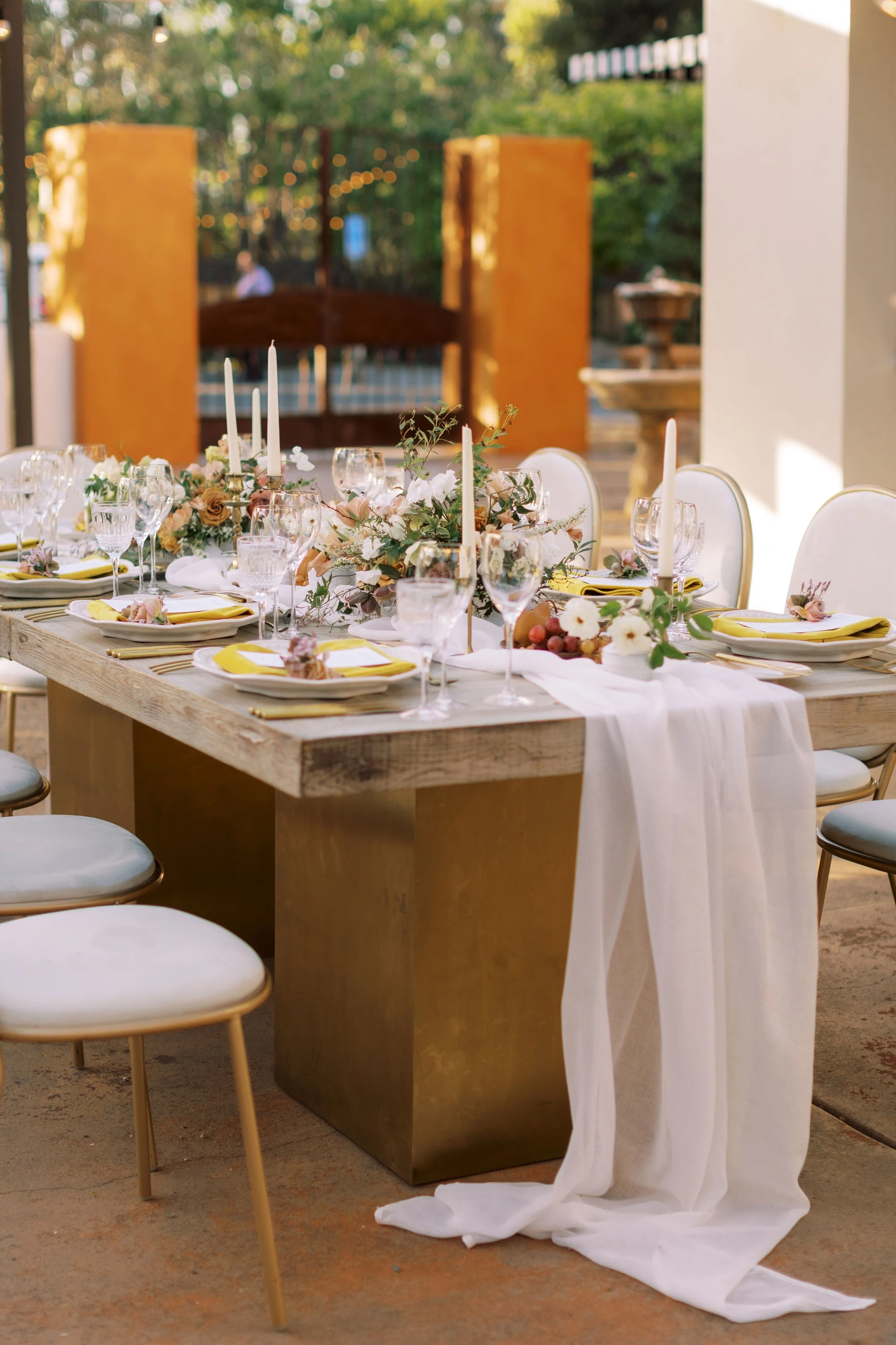 A long wooden table decorated with a floral arrangement, candles, and place settings, set outdoors on a sunny day with trees in the background.