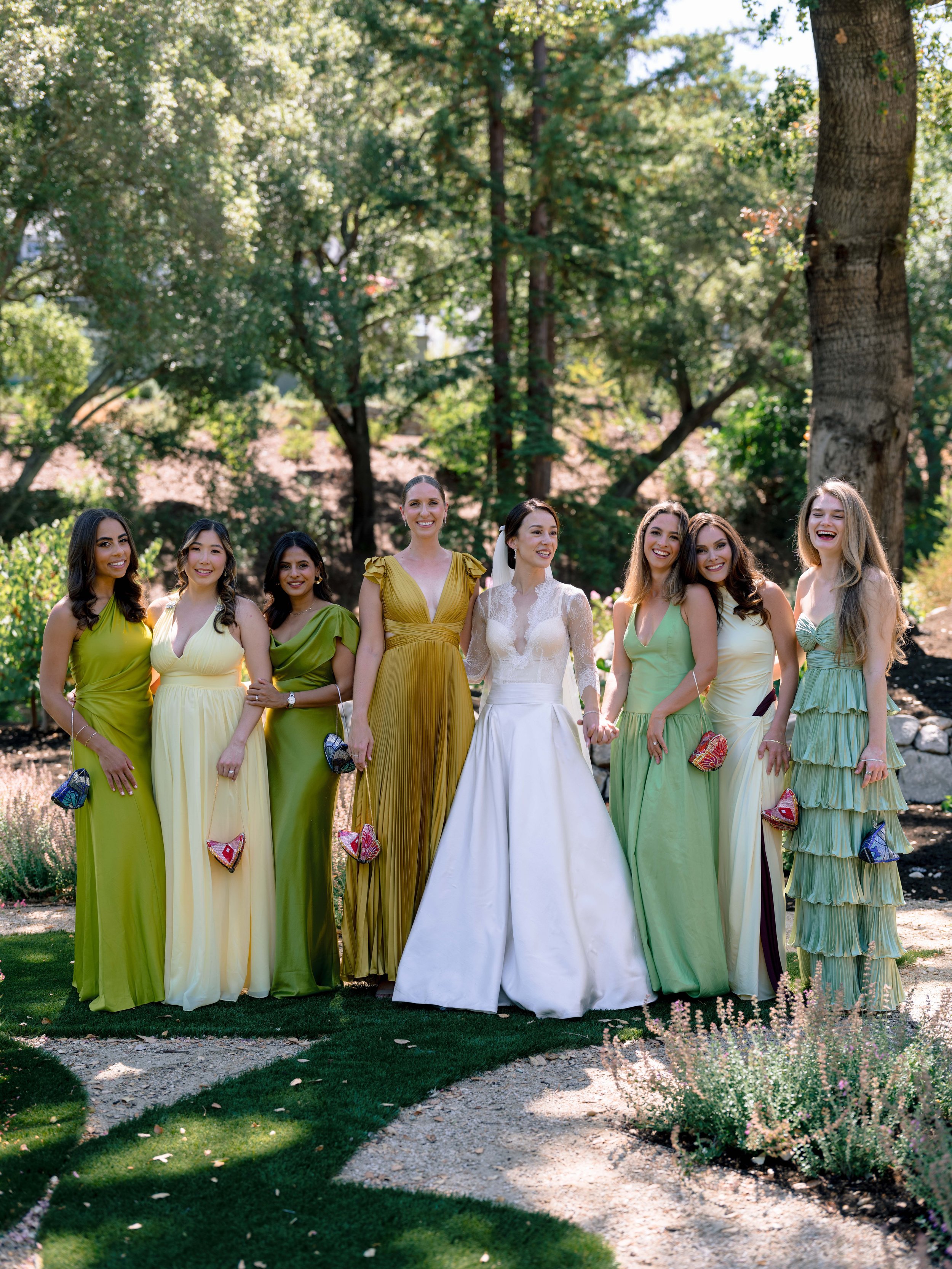 Group of women in colorful dresses standing outdoors in a garden surrounded by trees and plants.