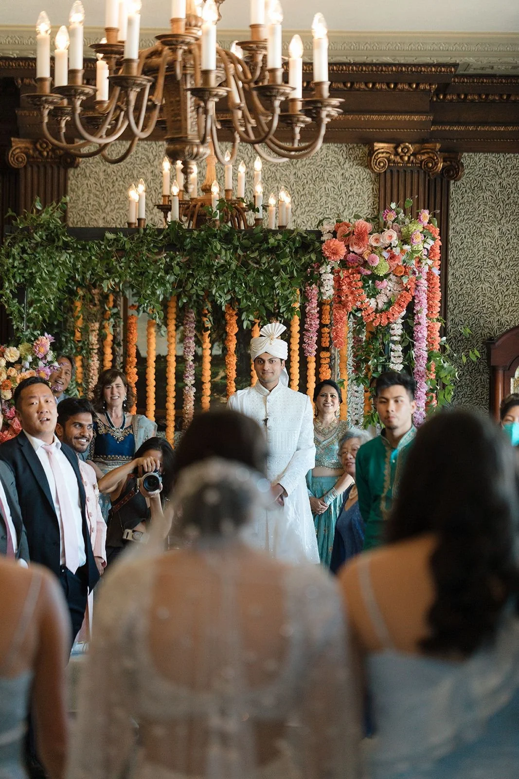 A diverse group of people celebrating a wedding indoors, with floral decorations and a chandelier above.