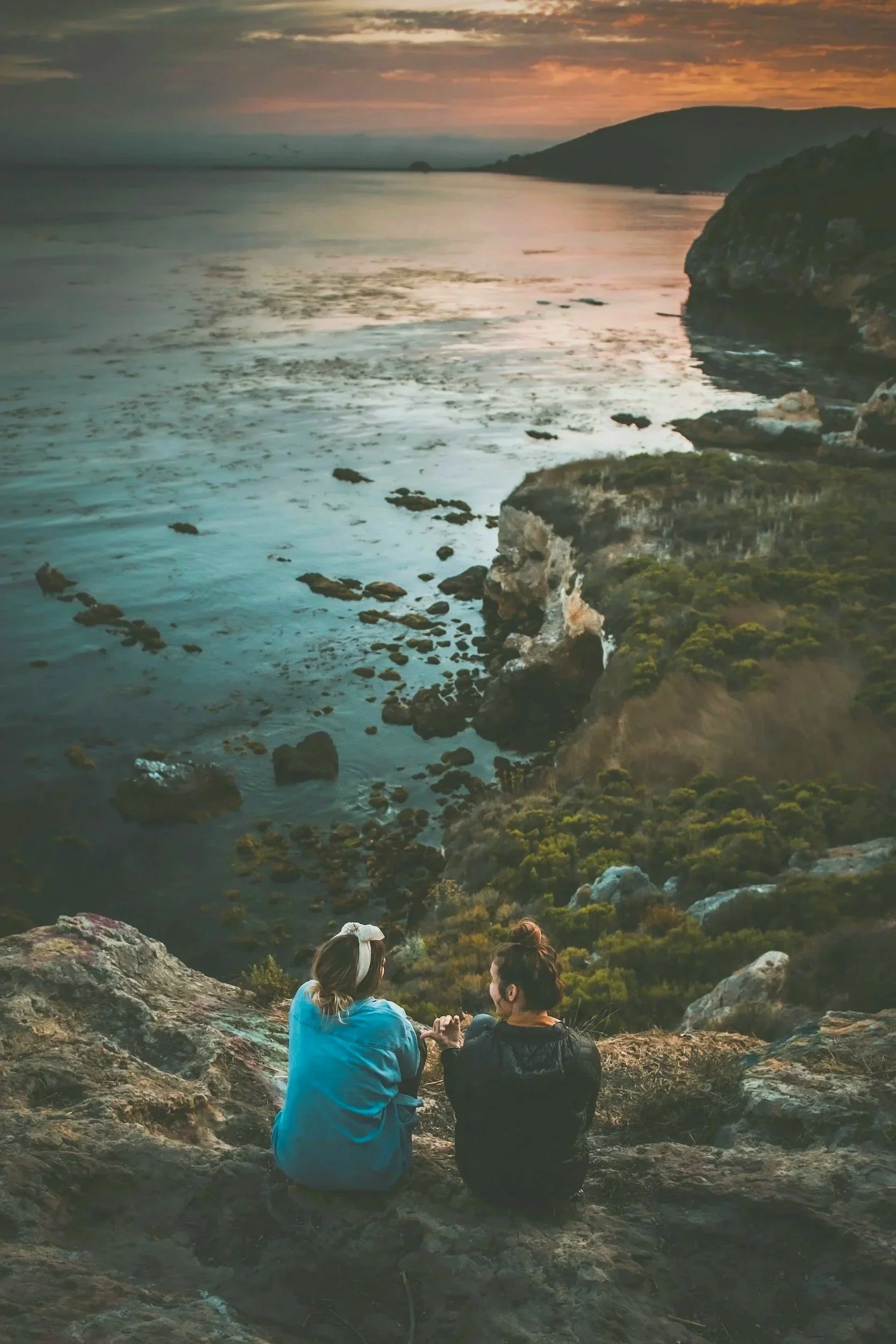 Two people sitting on a rocky cliff overlooking the ocean at sunset, with green hills in the distance.
