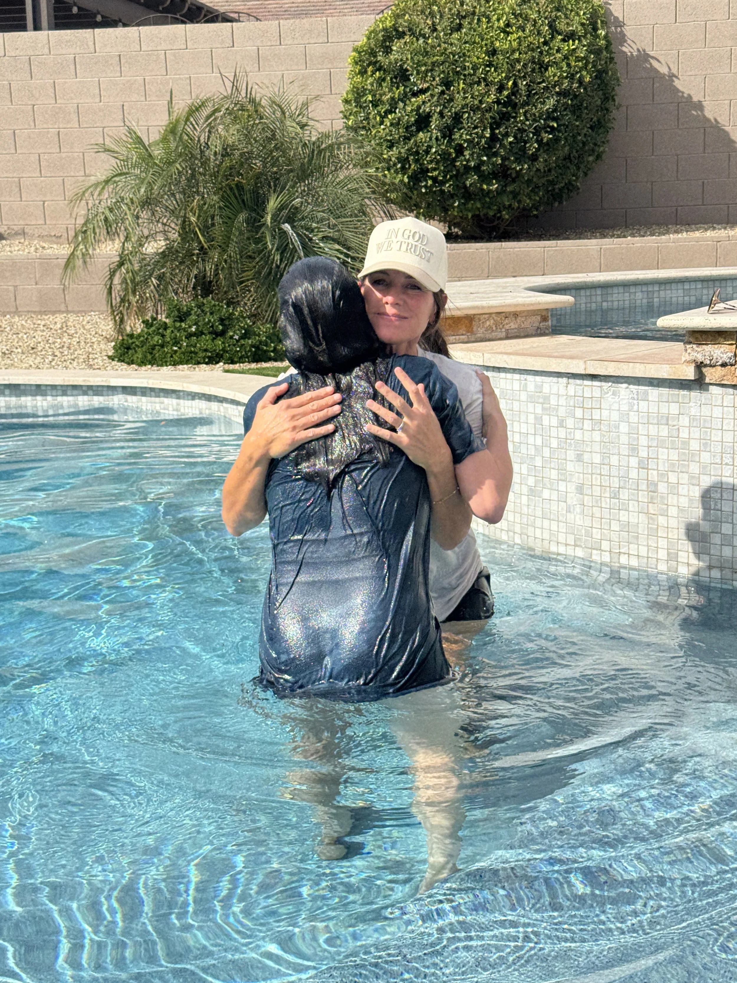 A woman hugging a person dressed as a black mermaid in a swimming pool. The woman is wearing a beige baseball cap with the words 'IN GOD WE TRUST' embroidered on it.