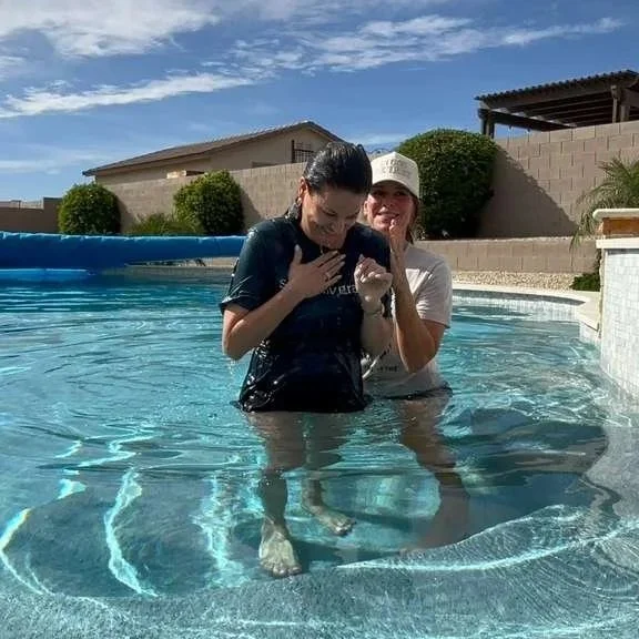 Two women are standing in a backyard swimming pool, one is wearing a black t-shirt and the other wearing a white cap and t-shirt, baptism