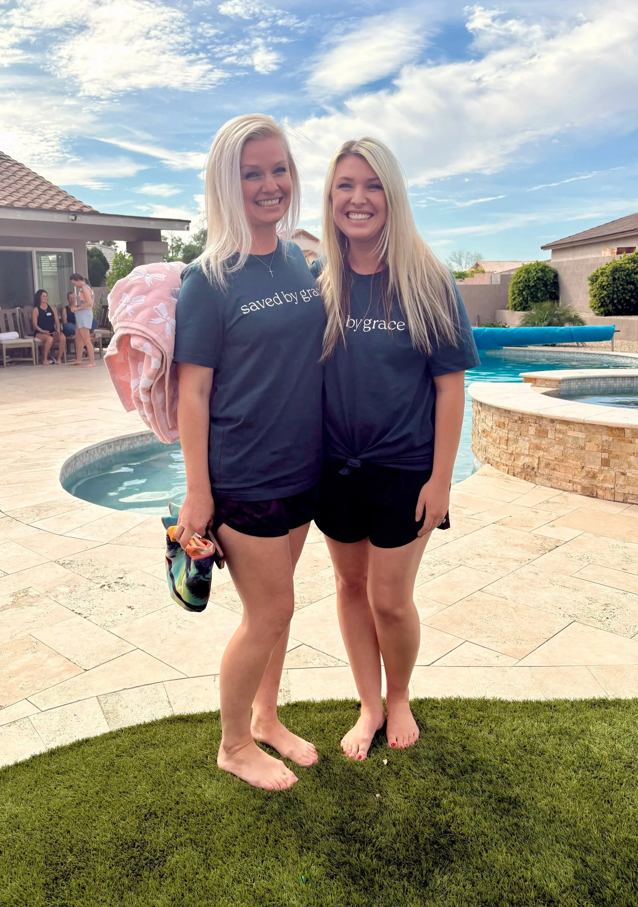 Two smiling young women with blonde hair, wearing matching navy blue T-shirts and shorts, barefoot, standing by the pool in a backyard on a sunny day.