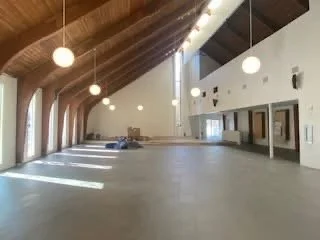 Image shows a large empty meeting area with round suspended lights, a gray floor, and white walls with oak beams and ceiling