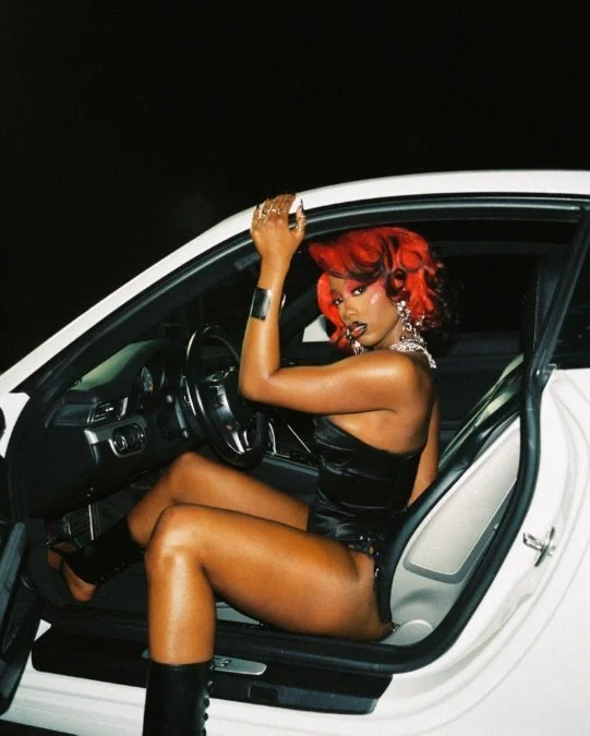 Flo Milli press photo by @fromakbar, artist with red curly hair sitting in a white luxury car wearing a black leather outfit with silver jewellery at night