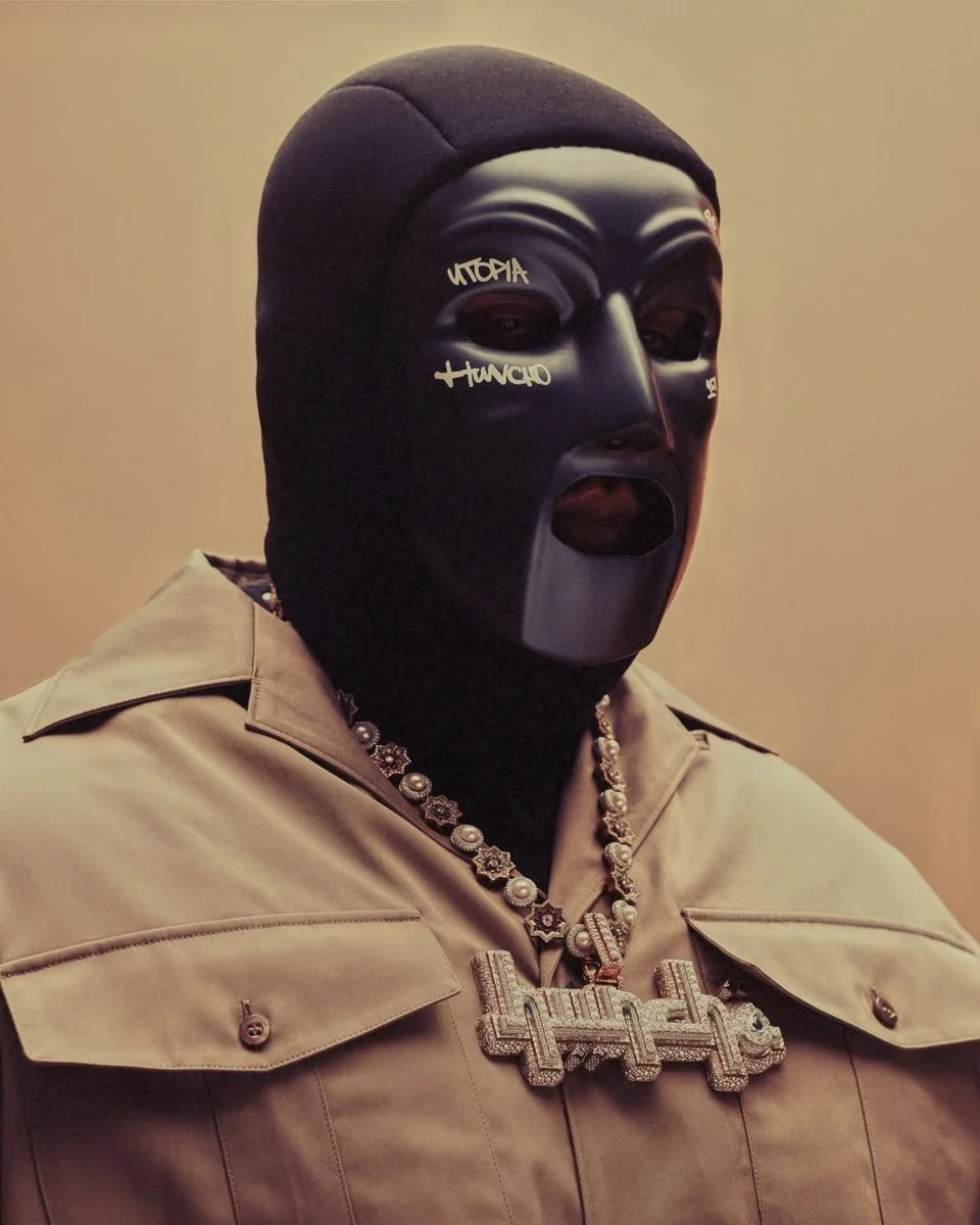 Press shot of rapper M Huncho wearing a black balaclava mask inscribed with Utopia Huncho, diamond chain and pendant, and a beige military-style jacket against a warm neutral background