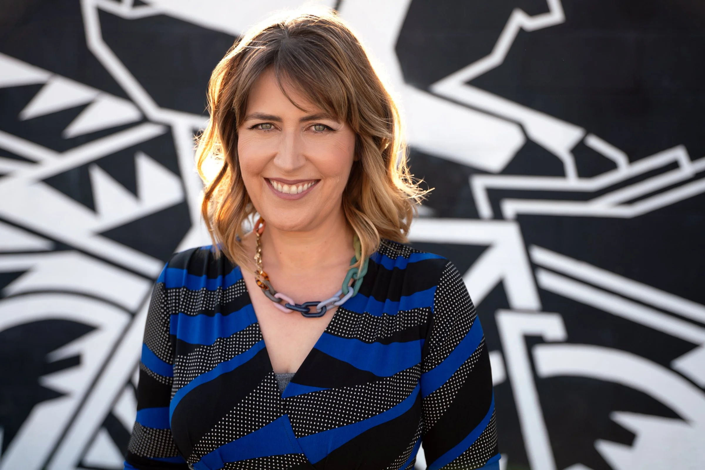 Close portrait of Darby Saxbe smiling in a blue patterned dress against a black and white graphic mural at The Spokein Los Angeles.