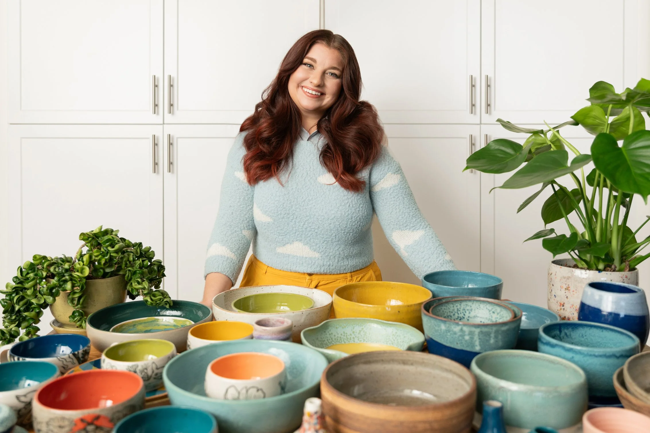 Ceramics business owner Leah standing behind a table filled with colorful handmade bowls in her kitchen, surrounded by houseplants.