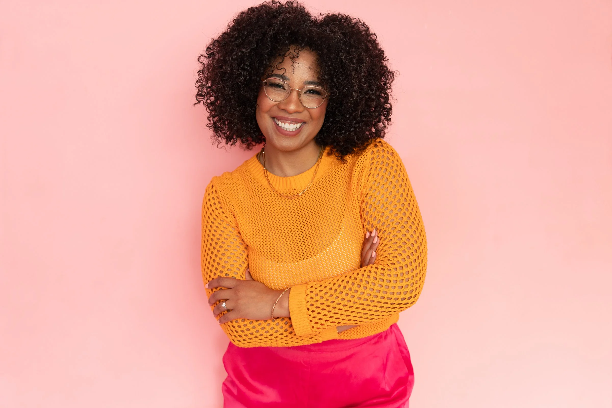 Dominique standing with arms crossed against a coral backdrop, wearing a mustard sweater and pink pants.