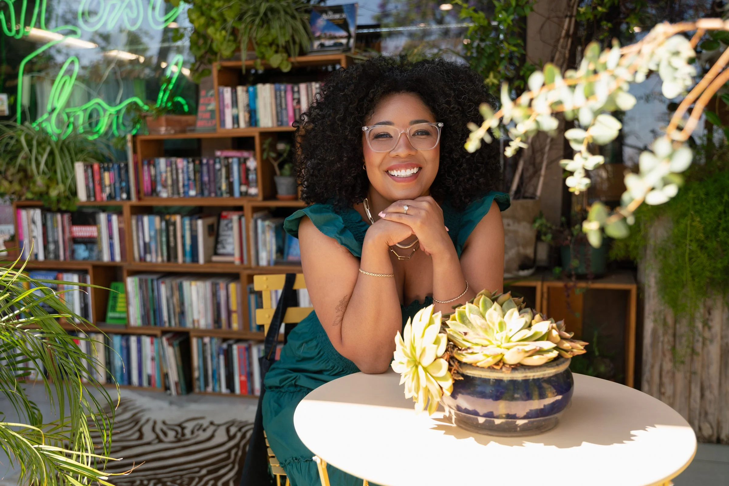 Dominique leaning on a table with flowers in front of a plant-filled bookstore, a green neon sign glowing behind her.