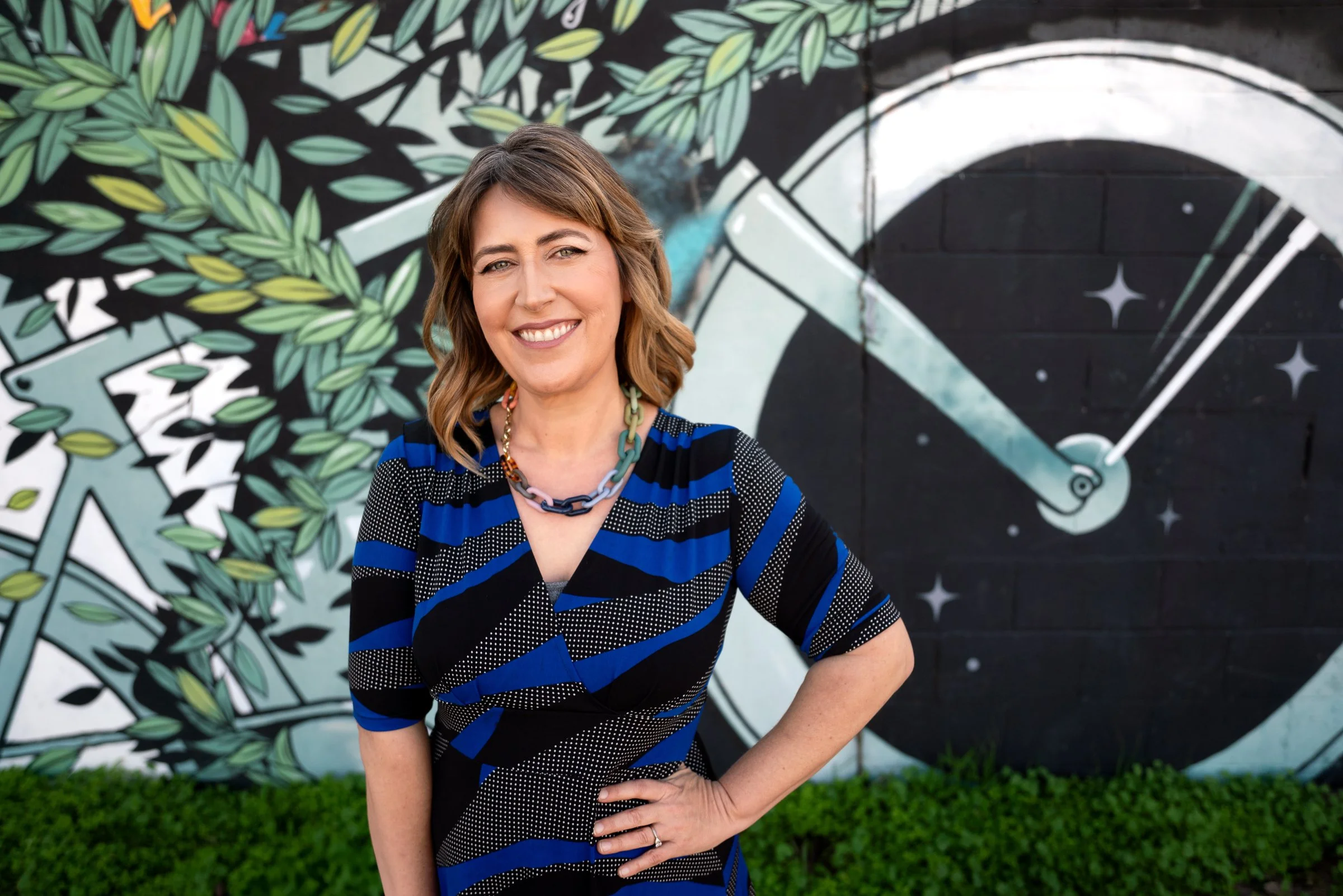 Darby Saxbe smiling with a hand on her hip in a blue striped top, in front of a leafy mural with a bicycle wheel graphic.