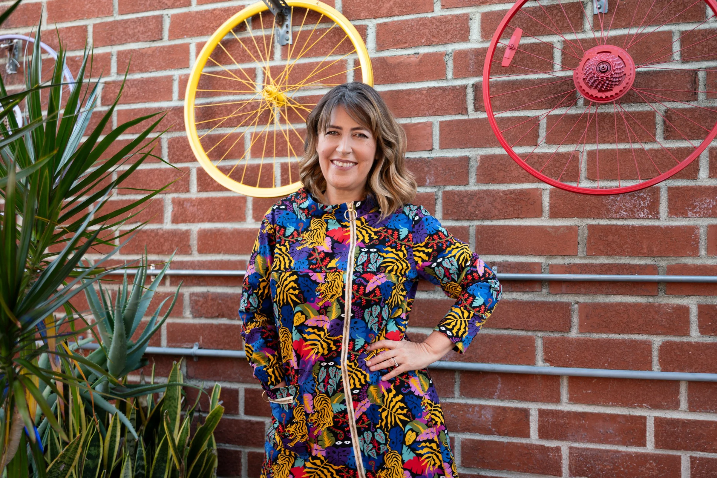 Darby Saxbe in a colorful Nooworks Jumper with a hand on her hip, standing against a brick wall with yellow and red bicycle wheels.