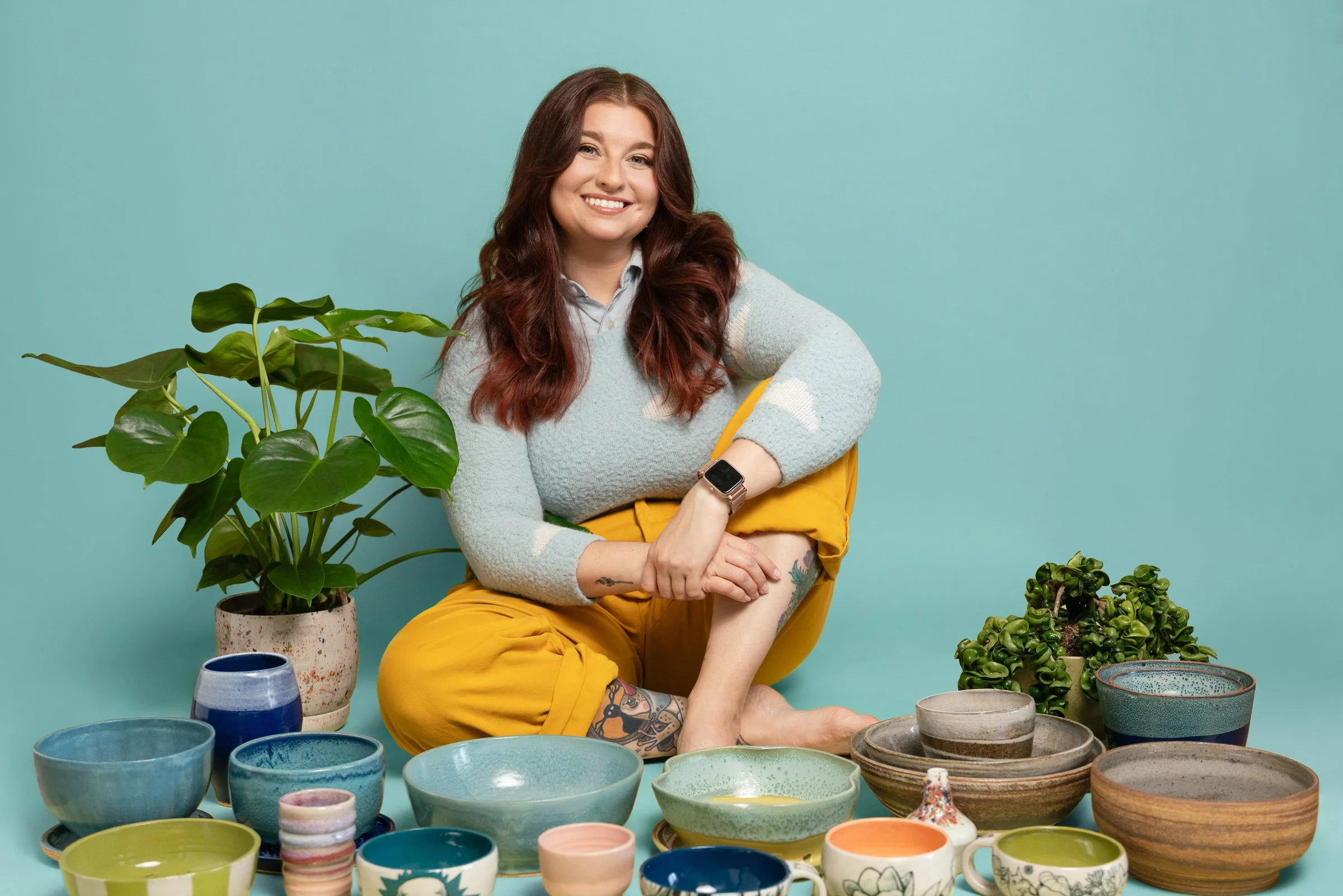 Leah seated on the floor surrounded by her handmade ceramic bowls and a potted plant, smiling against a teal backdrop.