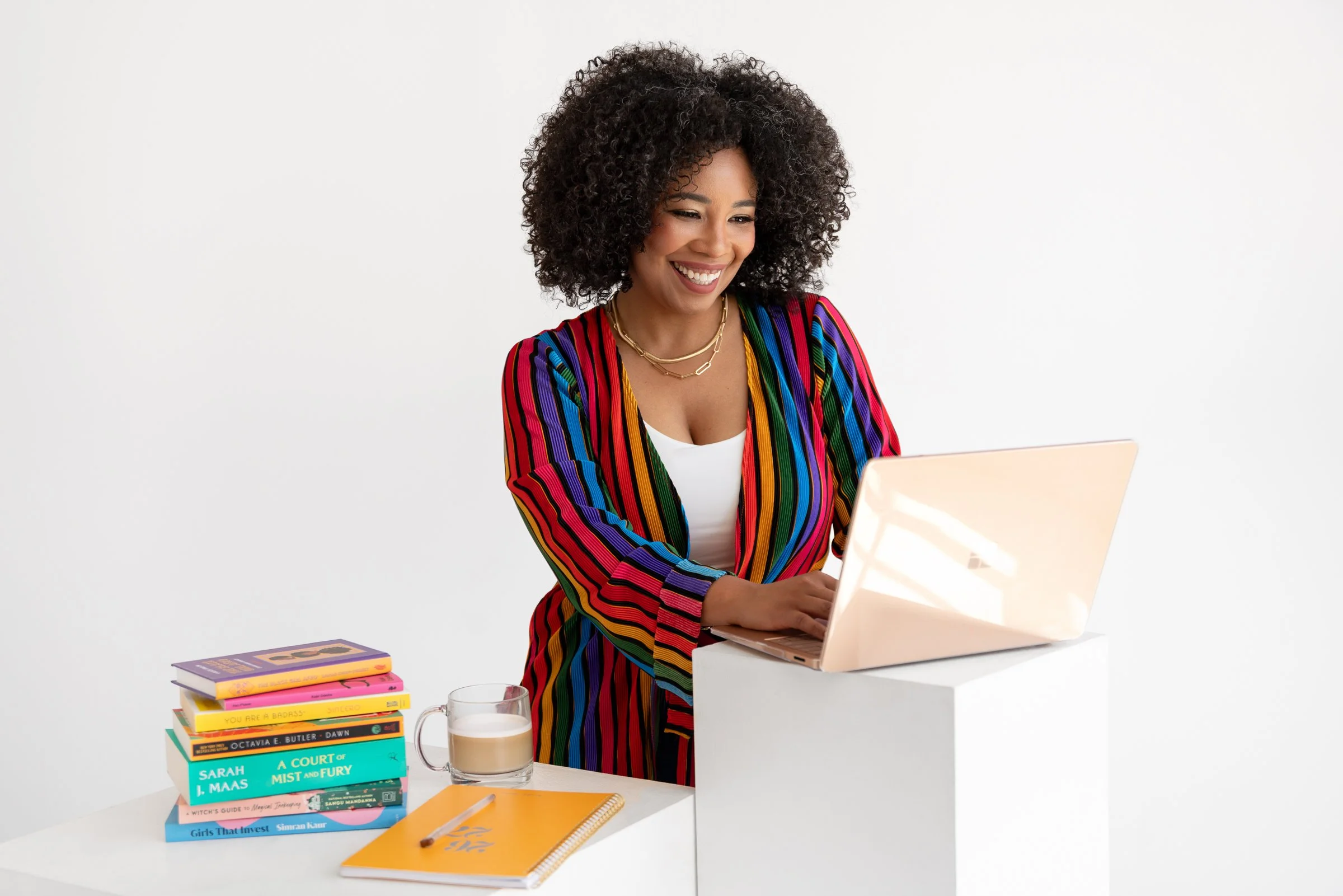 Dominique standing at a white "desk" with her laptop open, wearing a rainbow striped cardigan, books stacked beside her.