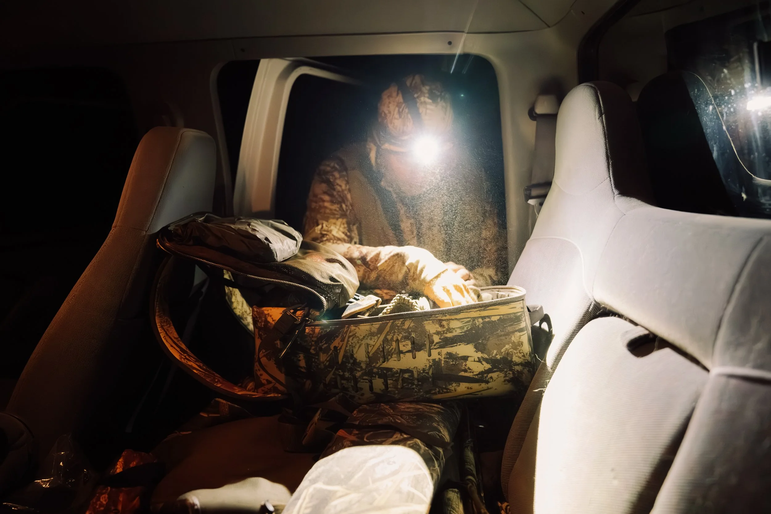 Person in a camouflage jacket standing outside a vehicle at night, taking a photo with a flash. The vehicle's interior shows a backpack on the back seat.