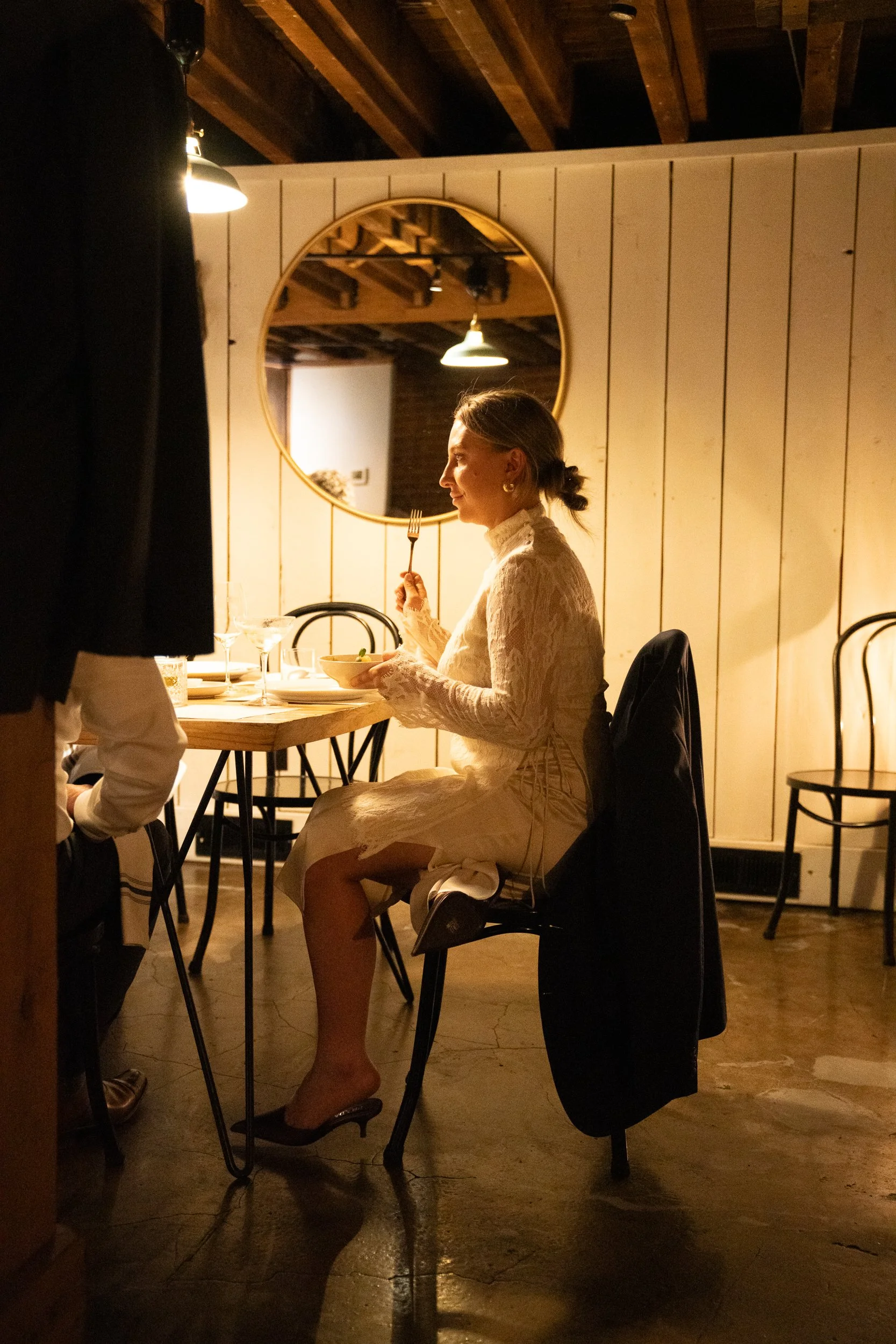 A woman in a white lace dress sitting at a dining table, holding a bowl and a fork, in a warmly lit restaurant with wooden walls and ceiling.