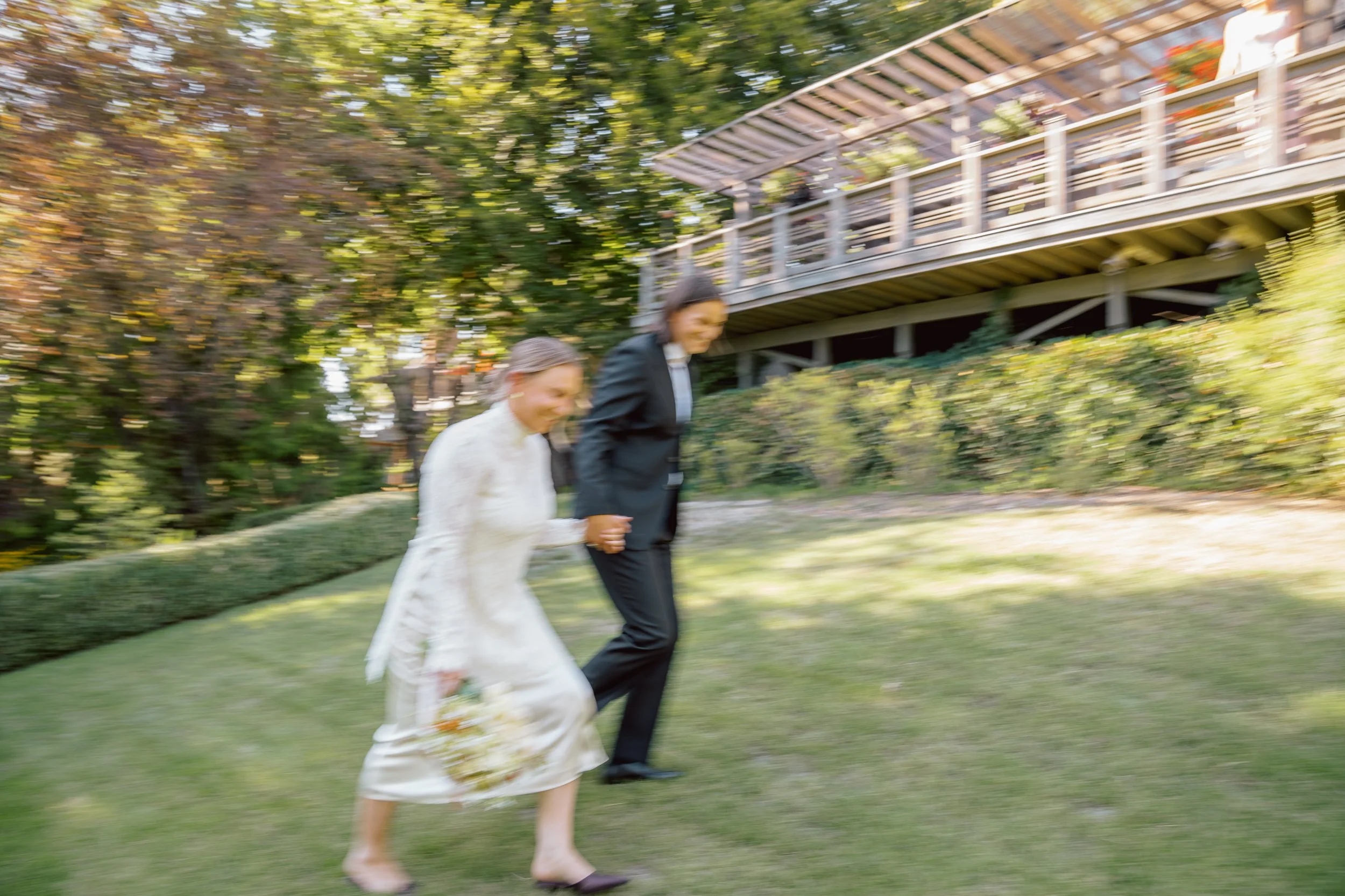 Two women, one in a white dress and the other in a black suit, walking hand-in-hand outdoors in a garden, slightly blurred, with trees and a wooden building in background.