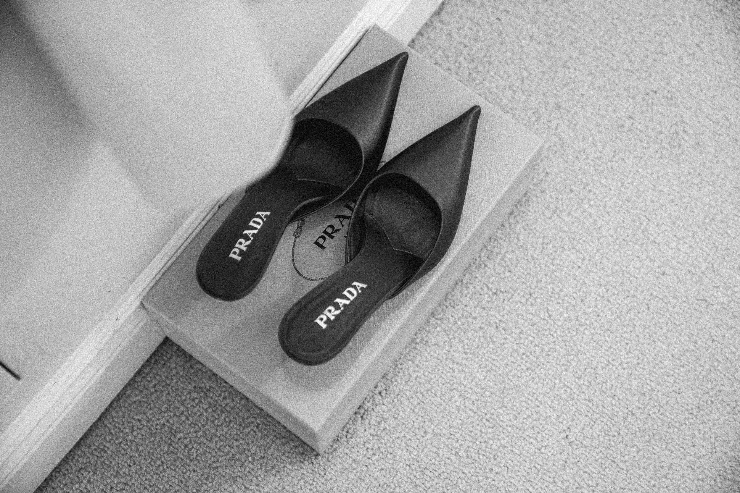 Black Prada high-heeled shoes with pointed toes on a shoe box on a carpeted floor.