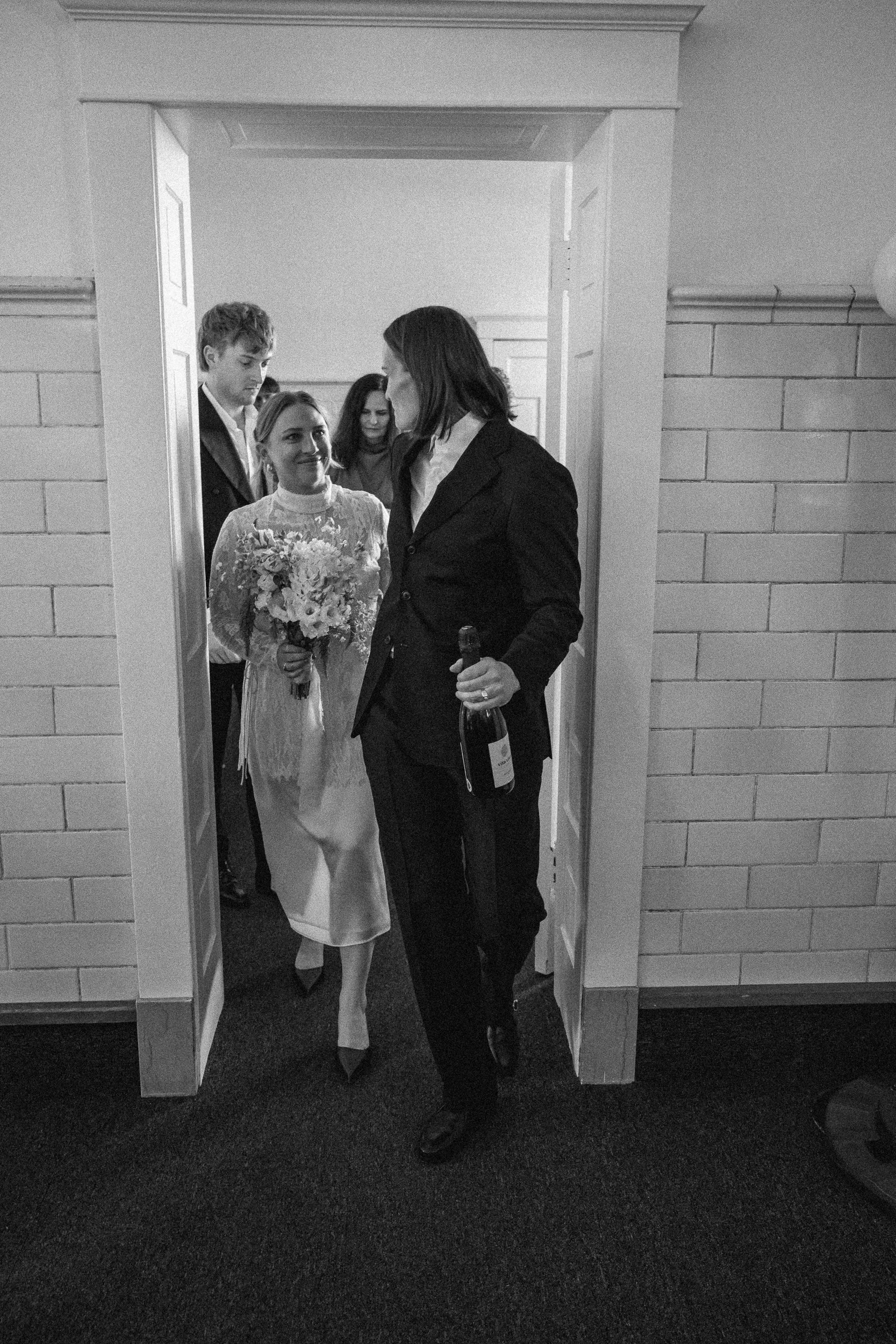 A group of people, including a woman in a wedding dress holding a bouquet, standing in a doorway having a conversation with a man holding a bottle. The scene appears to be at a wedding celebration.